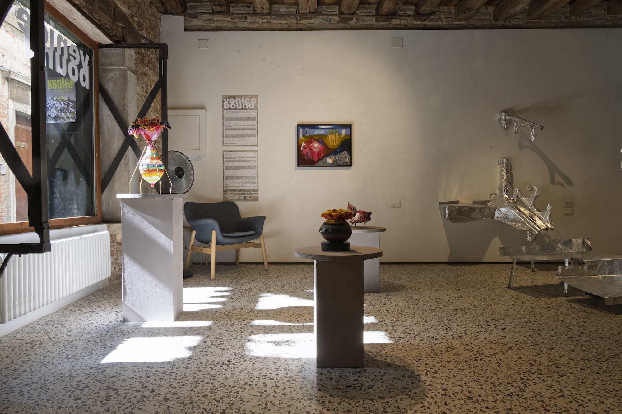 An art gallery interior with a central pedestal displaying a small sculpture, a framed painting on the back wall, and a metal installation on the right beside a grey chair and white partition.