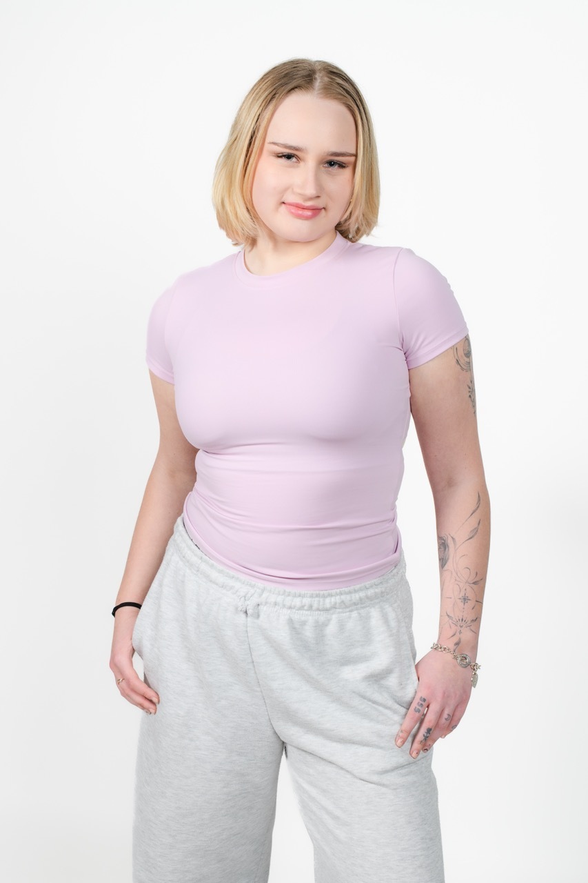 Woman with blonde bob wearing lilac fitted t-shirt and grey jogging bottoms, standing against white background with arm tattoos visible.