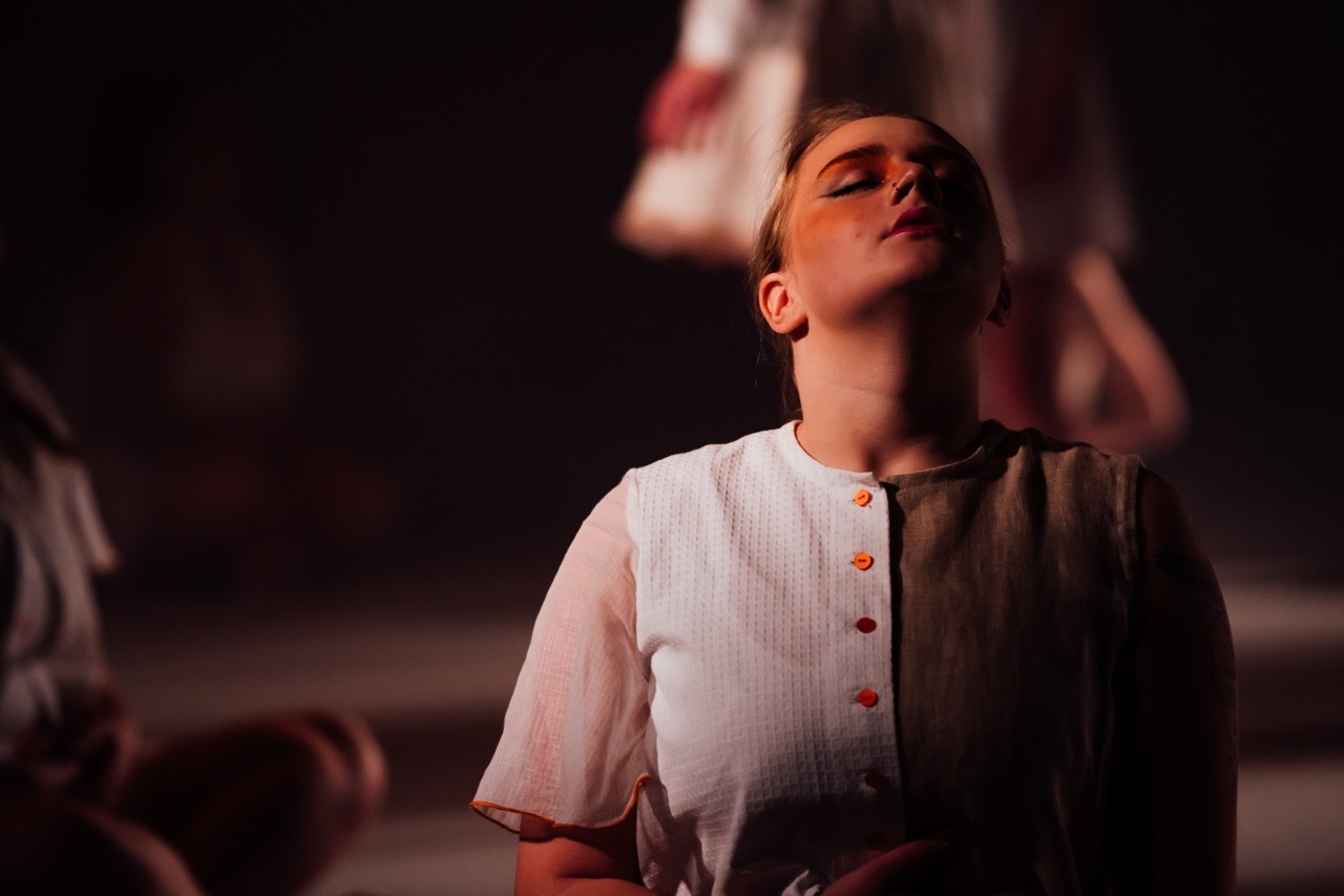 Woman in white button-up shirt with head tilted back, eyes closed, illuminated by warm orange lighting against dark background.