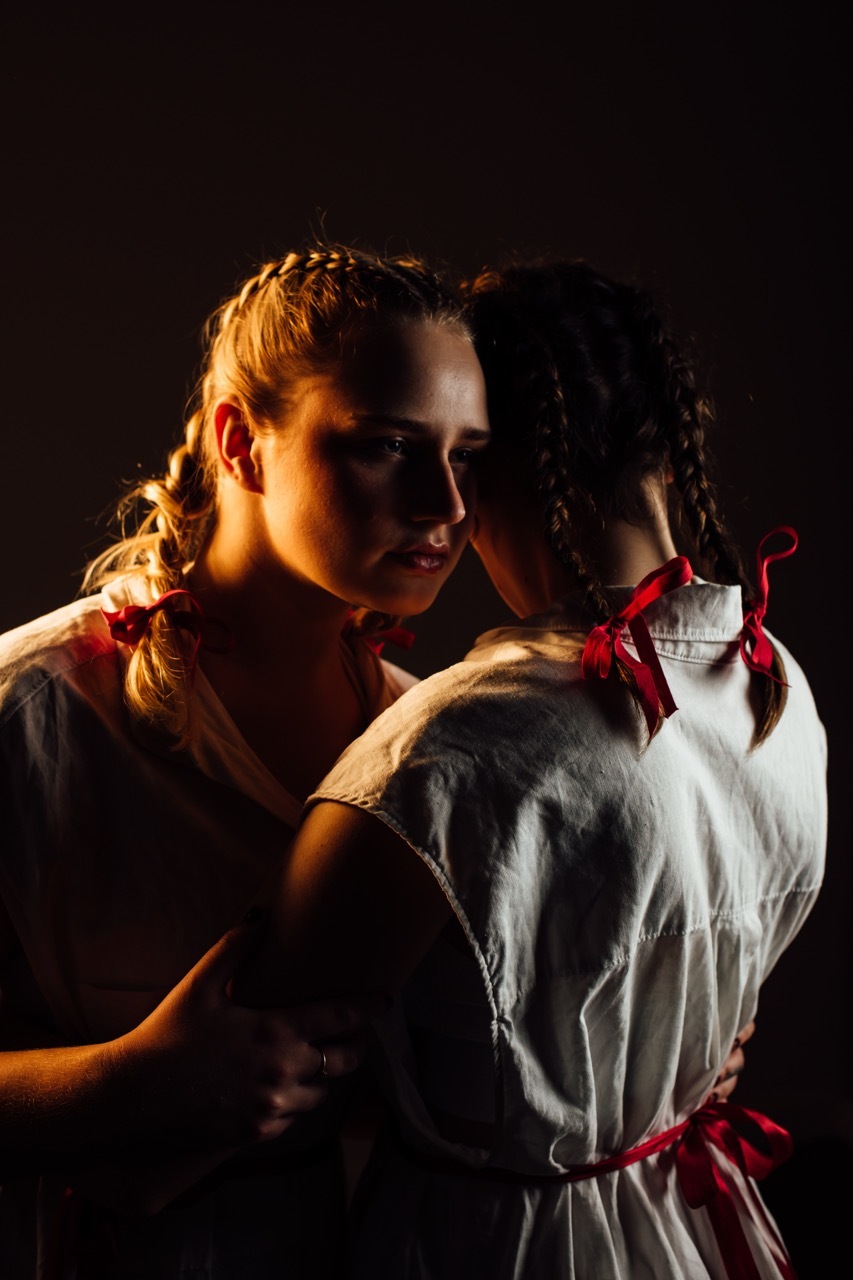 Two women embracing under dramatic stage lighting against dark background, one blonde in dark clothing, one brunette in white dress with red ribbons, warm lighting.