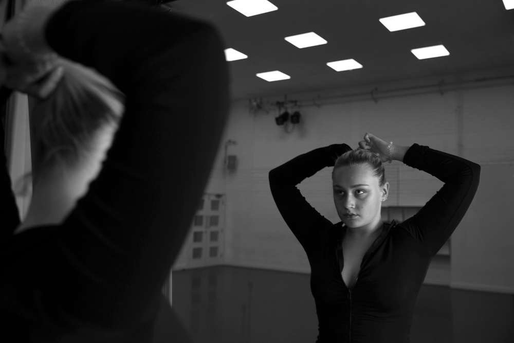 Woman with hands behind head in black top, reflected in mirror in gym or studio with square ceiling lights.