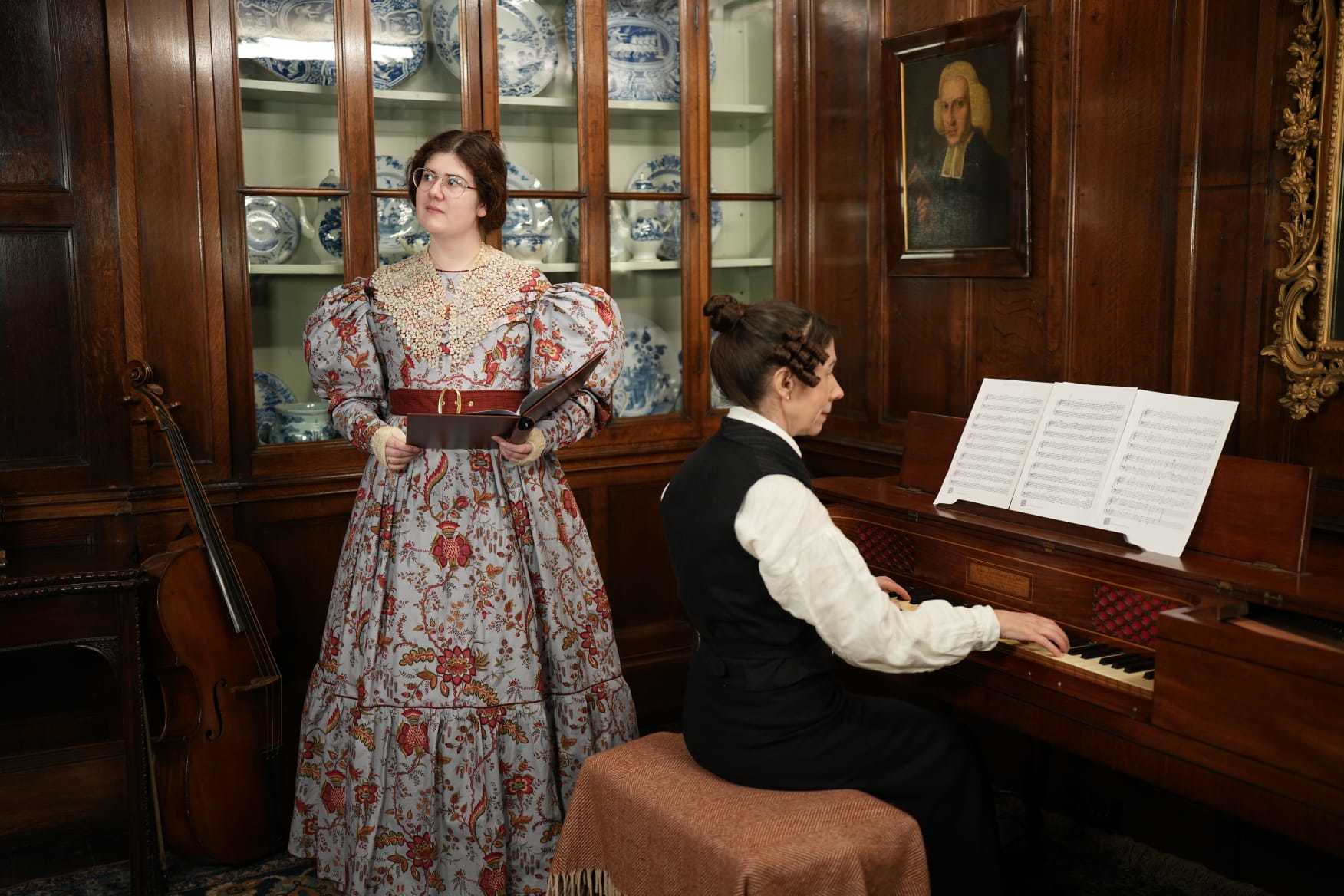 Singer V Joy in a Regency era costume holding a songbook, looking off to the left. A pianist in costume as Anne Lister sits at a piano and plays.