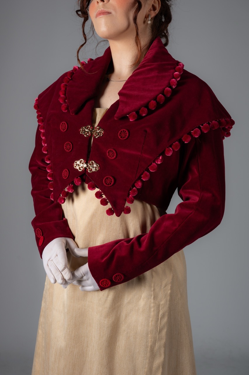 Woman wearing burgundy wool jacket with pom-pom trim and decorative buttons over cream dress, against grey background.