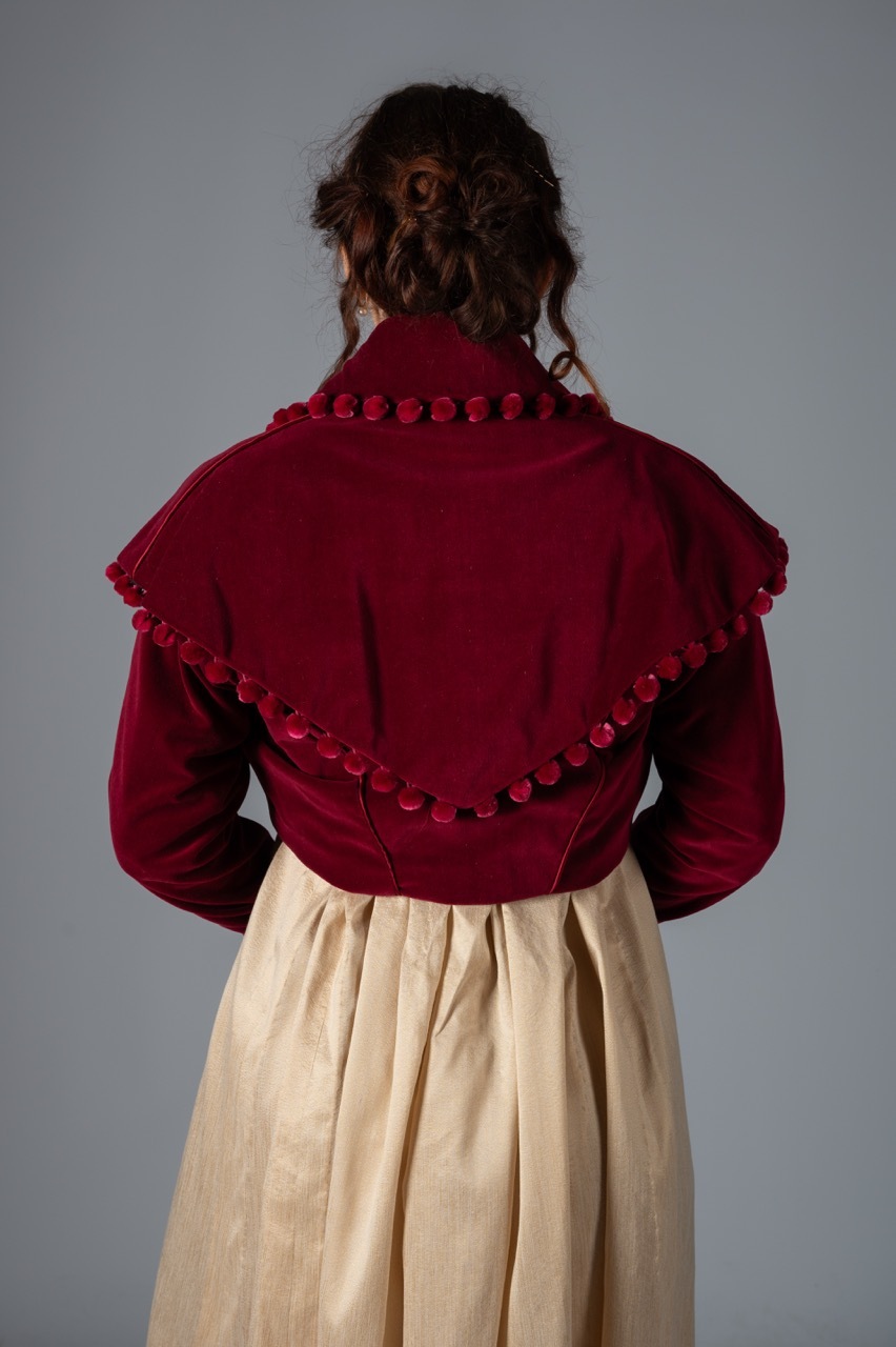 Woman from behind wearing burgundy velvet jacket with decorative buttons and cream-coloured skirt against grey background.