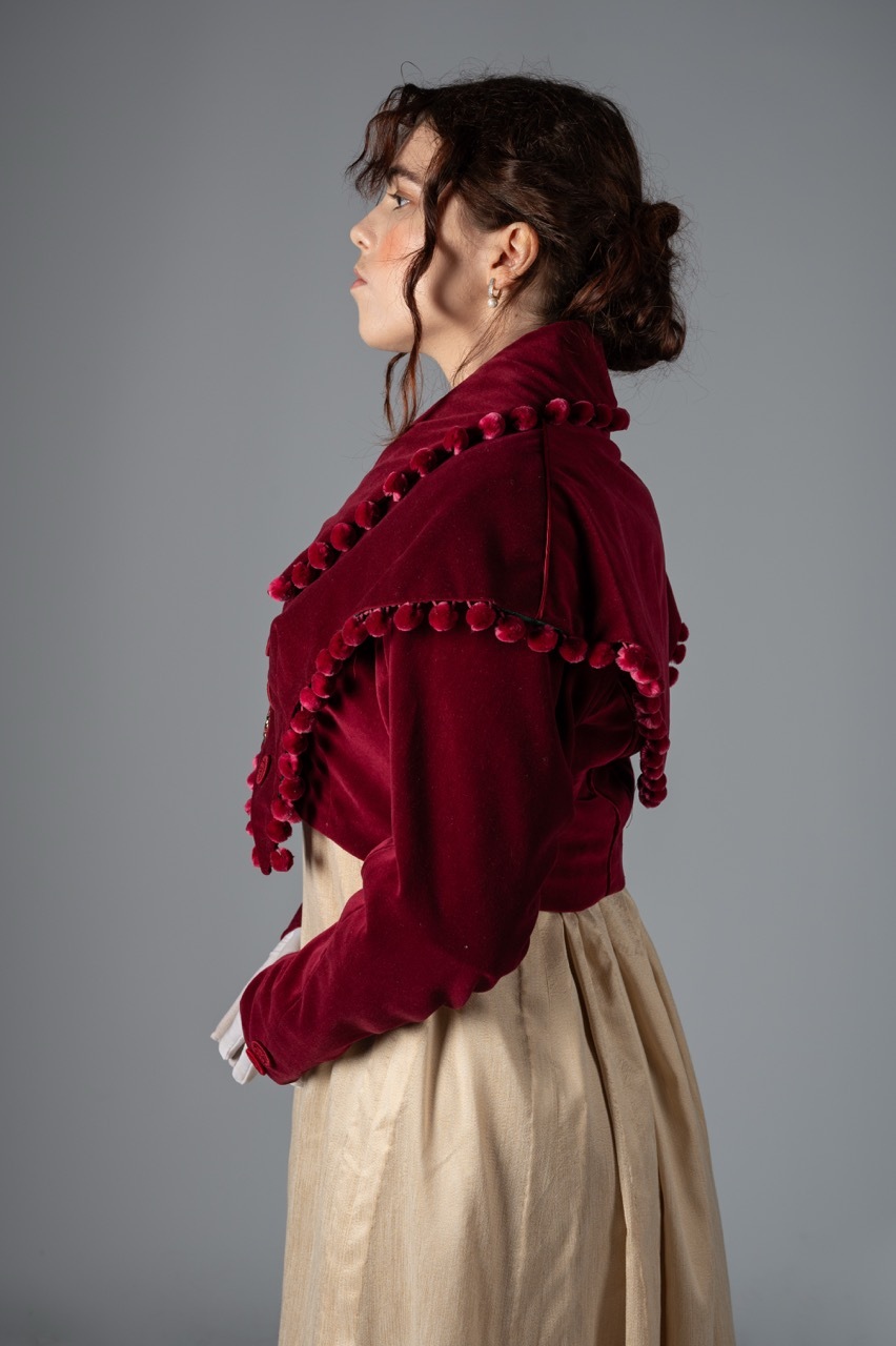 Woman in profile wearing burgundy velvet jacket with pom-pom trim and cream-coloured skirt against grey backdrop.