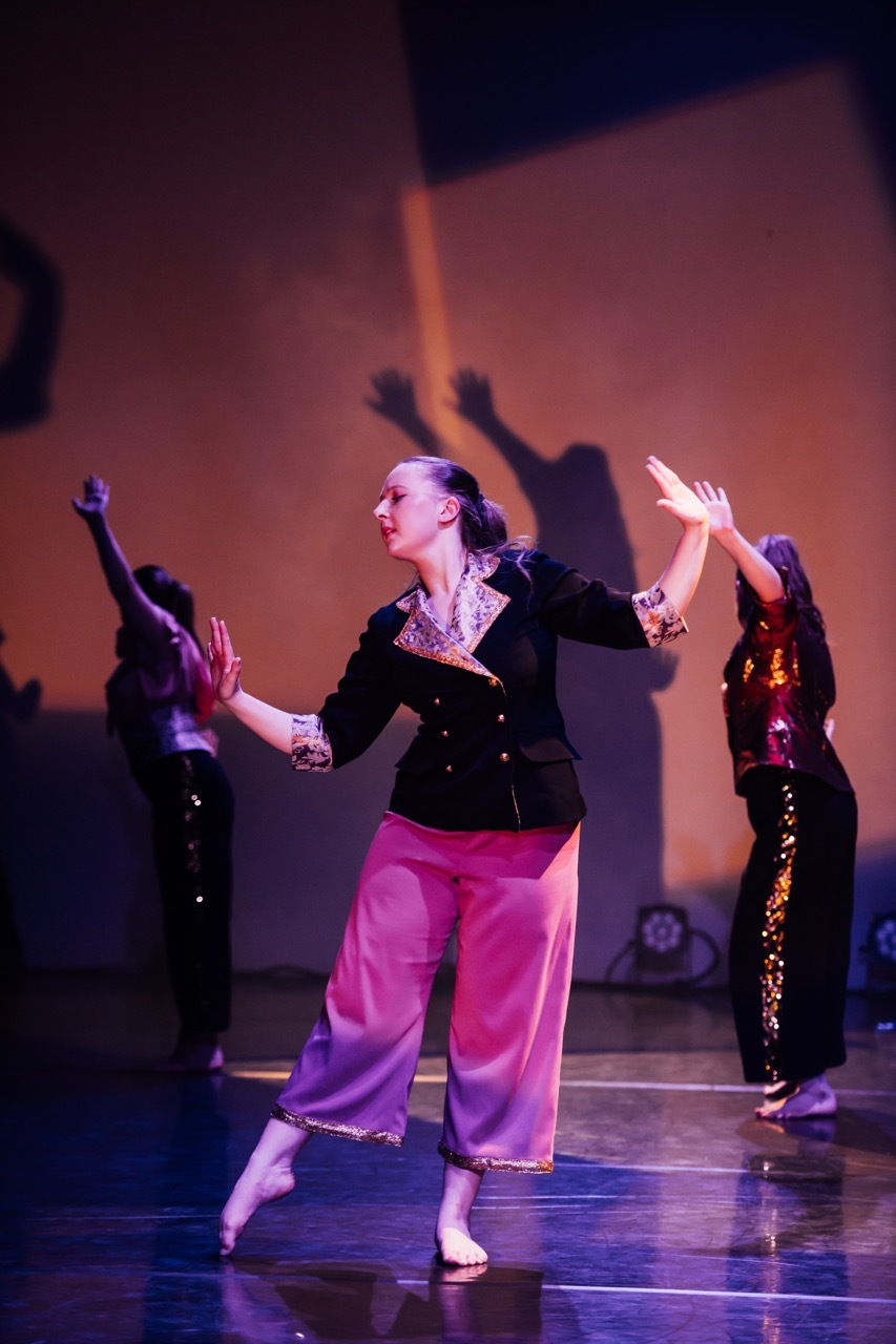 Three dancers on stage with arms raised. Centre performer wears black top and bright pink trousers. Warm orange lighting on backdrop.