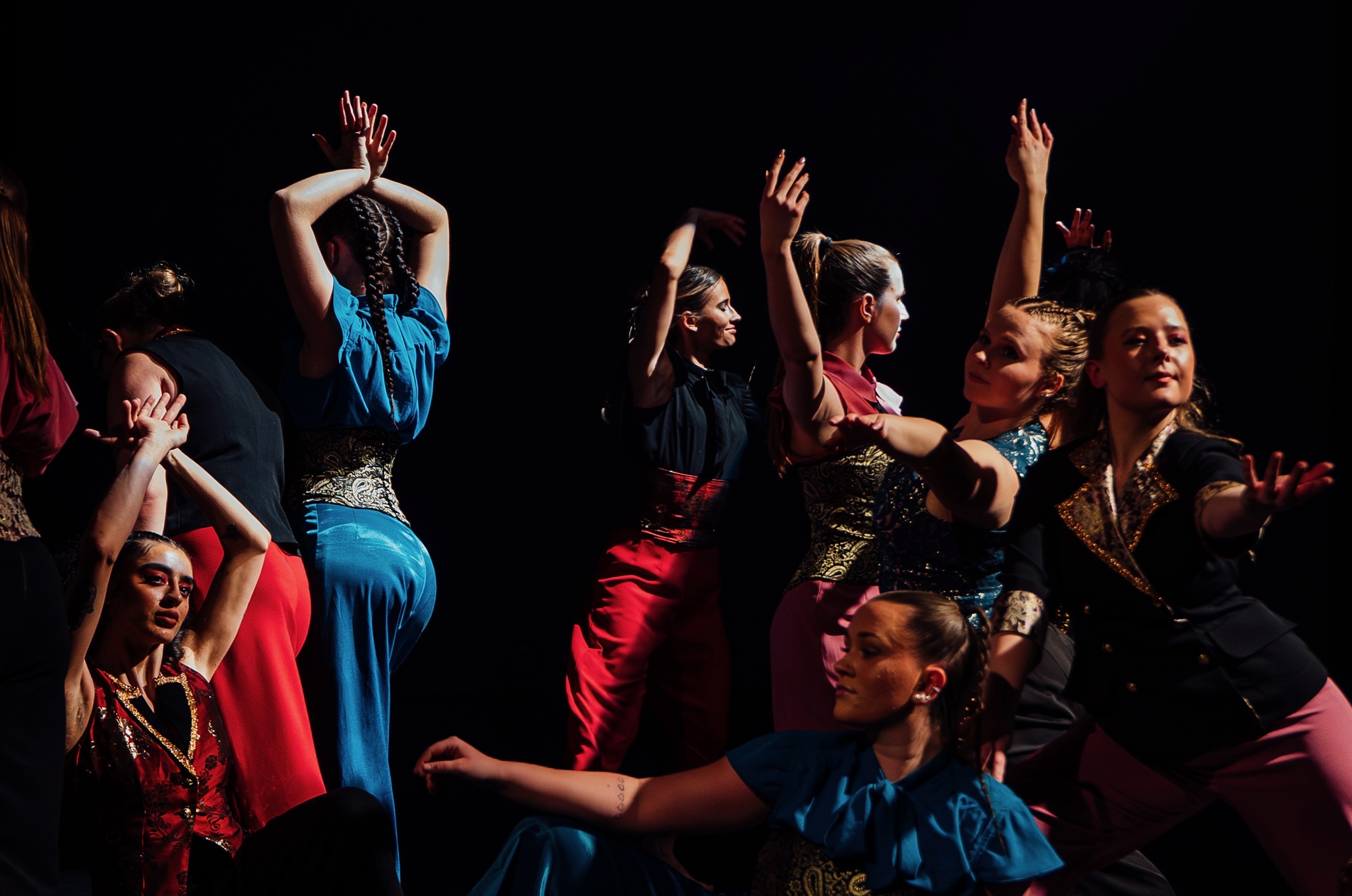 Group of performers in colourful costumes with raised arms against black background, illuminated by stage lighting.