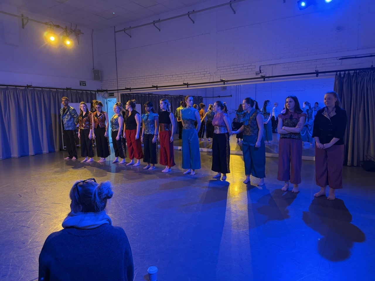 Group of people standing in line on wooden floor in hall with blue and yellow stage lighting, grey curtains visible on walls.