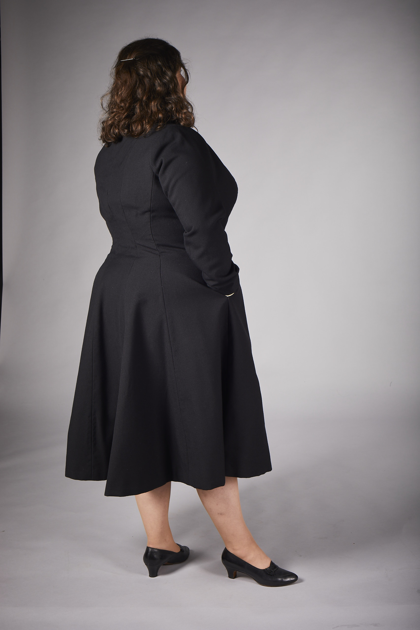 View of the back of a long black wool coat, paired with small black heels. The model’s hands are in the pockets of the coat.