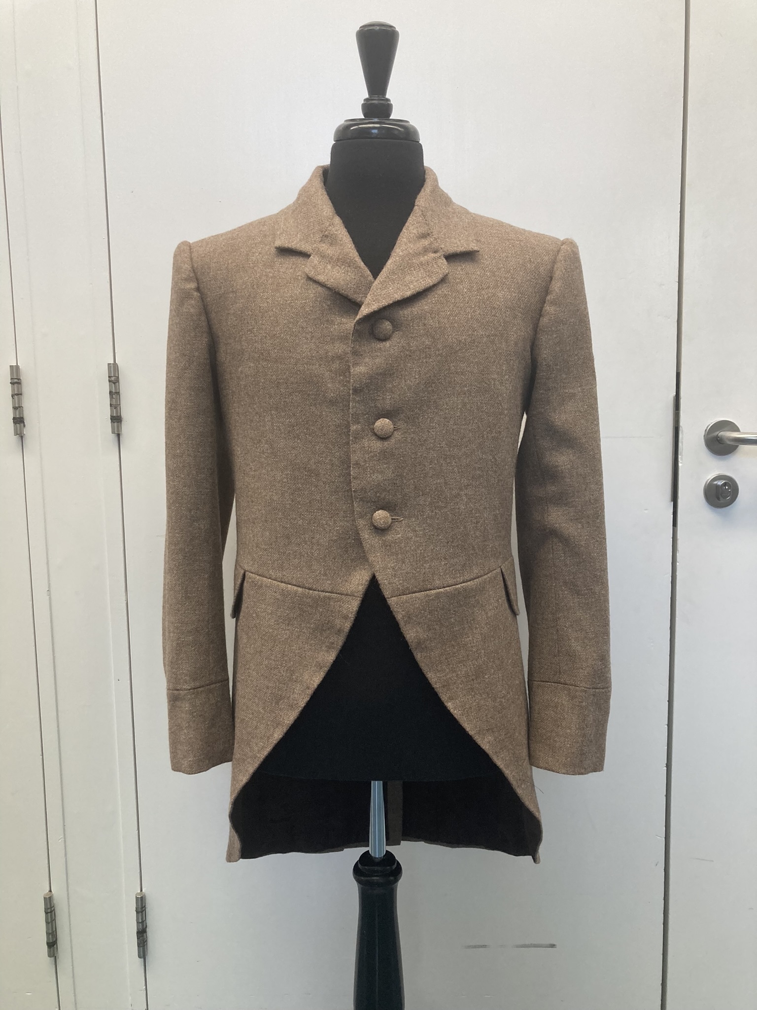 Tan wool tailcoat with four buttons displayed on black dress form against white mirrored wardrobe doors.