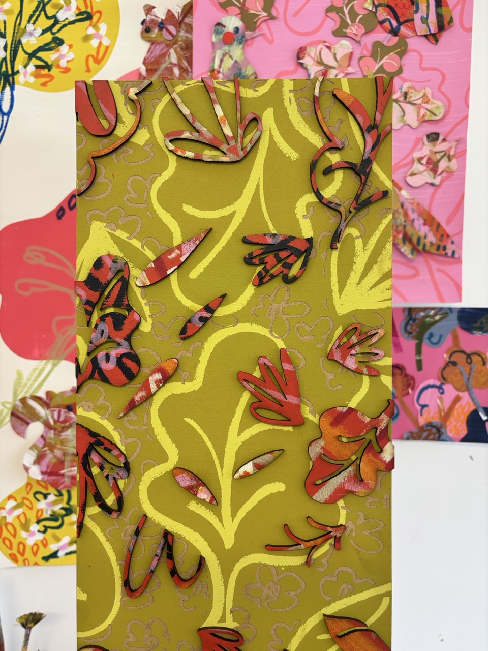A vertical decorative panel featuring a vibrant green background with intricate yellow and red floral designs. Behind it are blurred images of pink and patterned artworks, creating a layered visual effect.