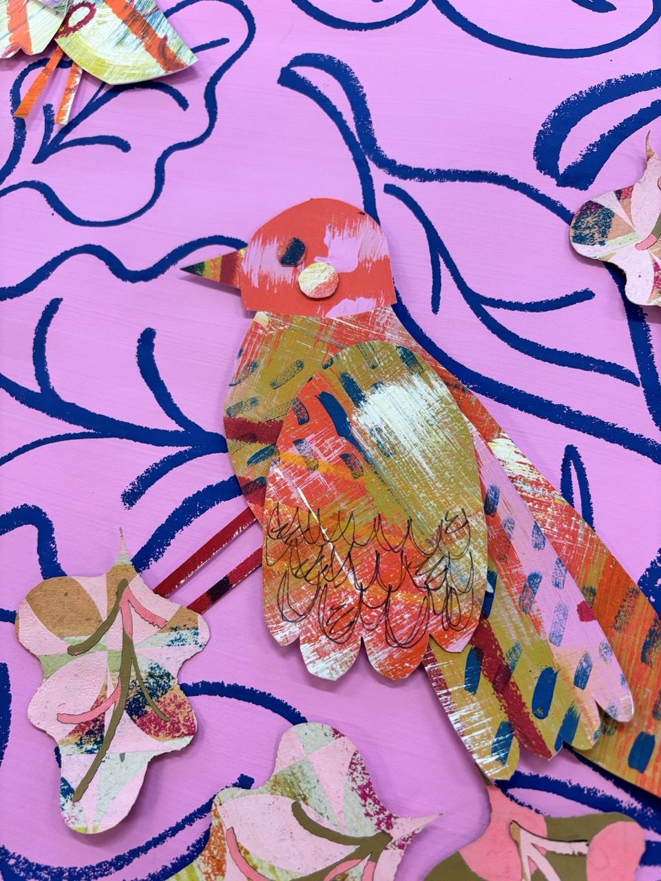 A brightly coloured bird illustration made of cut paper, featuring a pink body with red and orange patterns. The background is painted in pink with blue floral motifs, and additional cut paper flowers are visible around the bird.