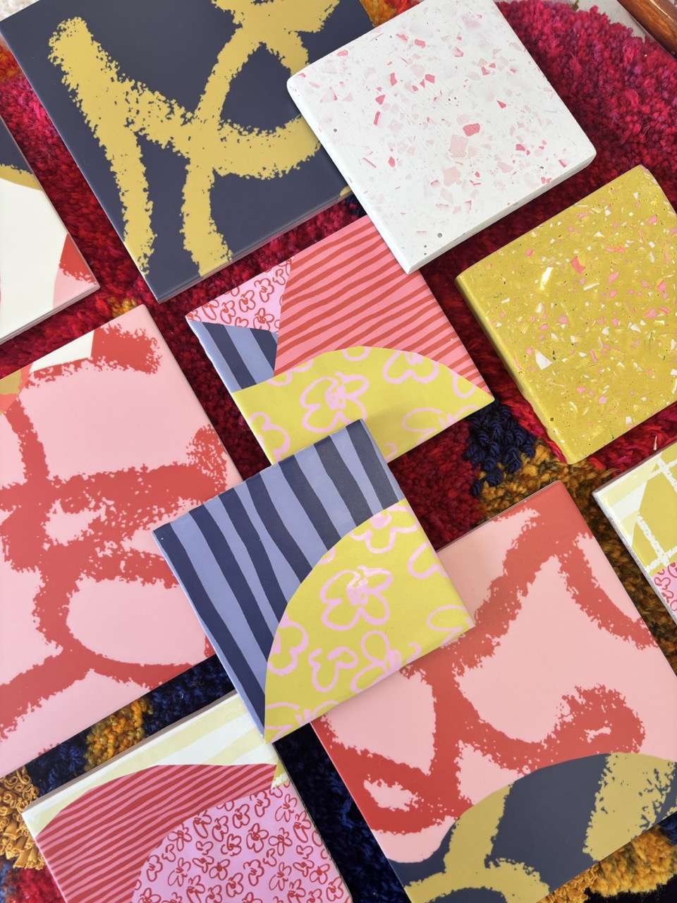 A collection of square ceramic tiles featuring various abstract patterns and colours, including pinks, yellows, and whites. The tiles are arranged on a textured surface with a red and yellow backdrop, highlighting their decorative designs.