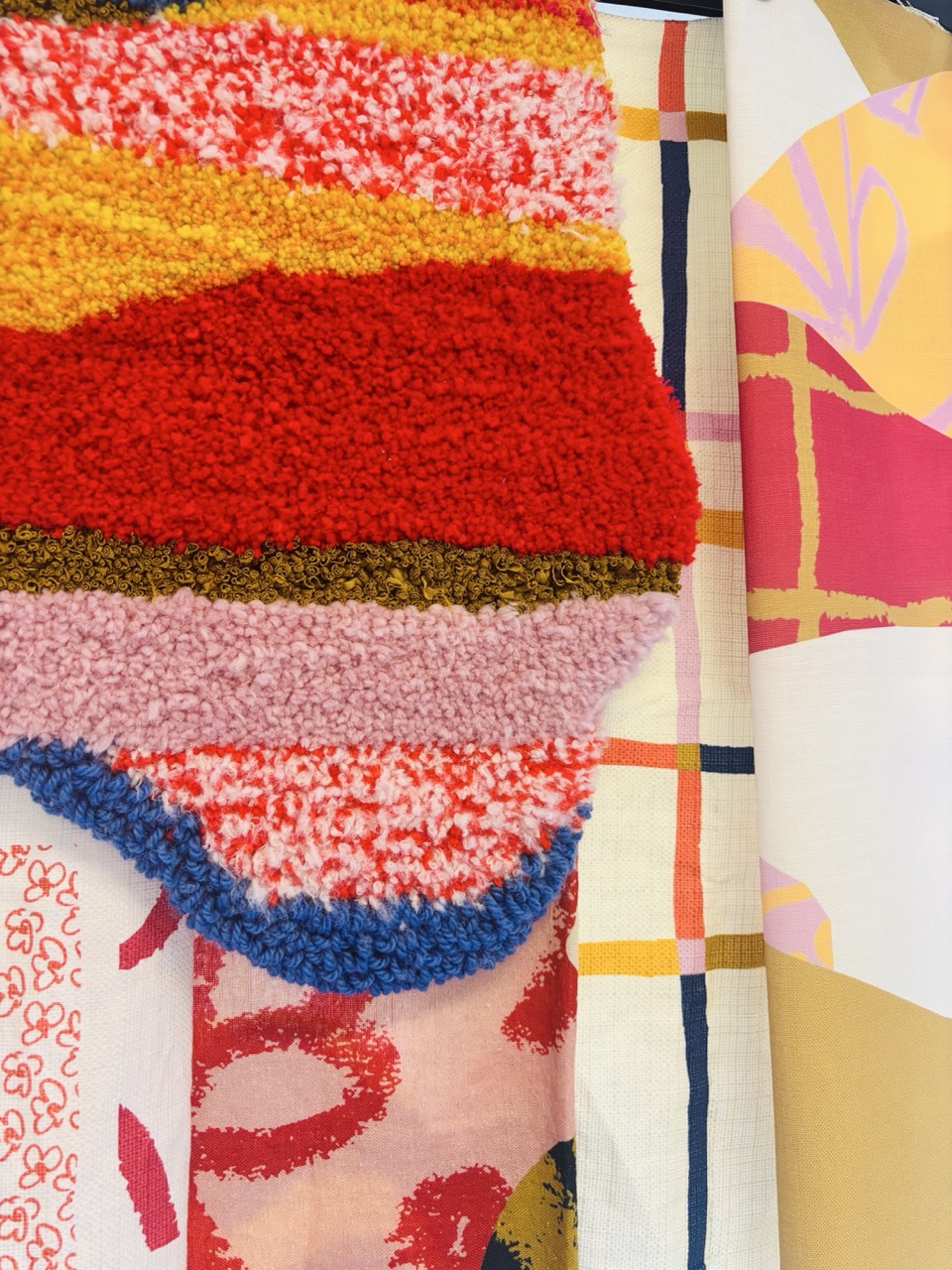 A collection of patterned fabrics hung together. The top features a striped rug with bright colours, including red, yellow, and pink, beneath which are various textile designs, including floral and geometric patterns in shades of pink, cream, and brown.