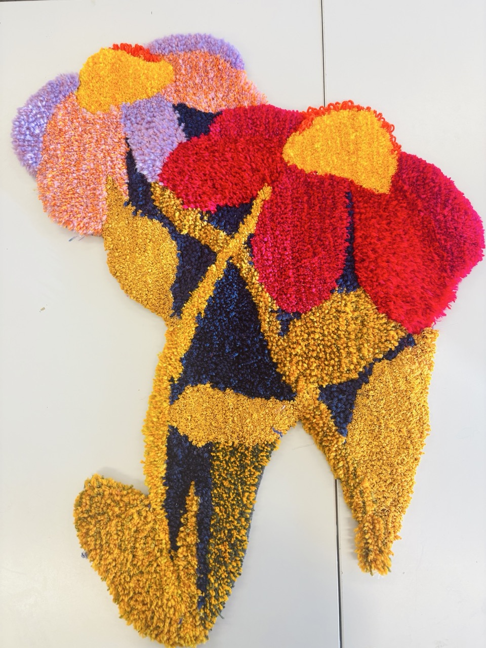 A colourful rug shaped like a silhouette with abstract floral designs. The rug features large flowers in red, pink, and purple, set against a yellow and dark blue background, creating a striking visual pattern.
