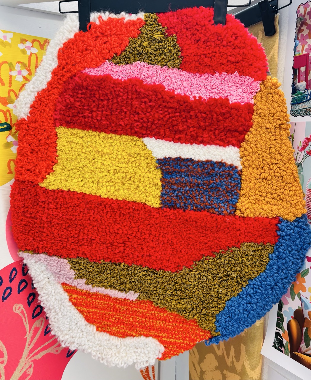 A textured wall hanging featuring a mix of vibrant coloured yarns in a geometric pattern. The design includes shades of red, yellow, blue, pink, and green, arranged in stripes and sections, creating a bold, artistic appearance.