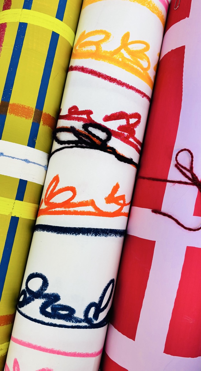 Three rolled-up sheets of patterned paper are displayed. The colorful designs include yellow and blue striped patterns, red and pink sections with abstract lines, and orange accents featuring handwritten-like motifs on a white background.
