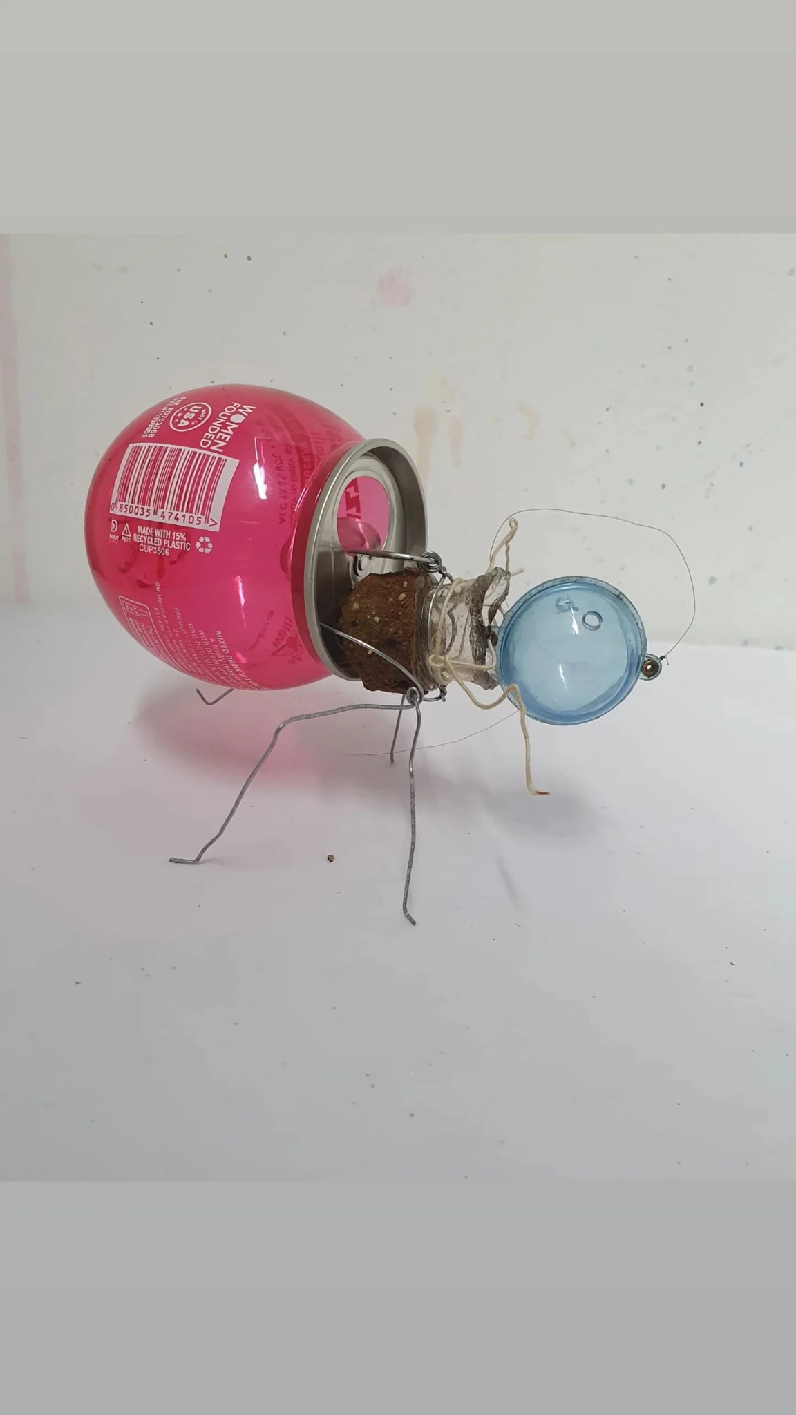 A small handmade robot assembly made from a pink can and a blue plastic cup, with wires and thin metal legs on a white surface.