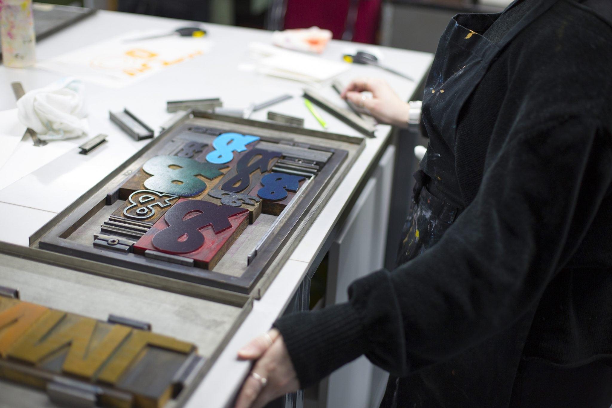 A person standing by a screen printer with an assortment of ampersand shapes inside it.
