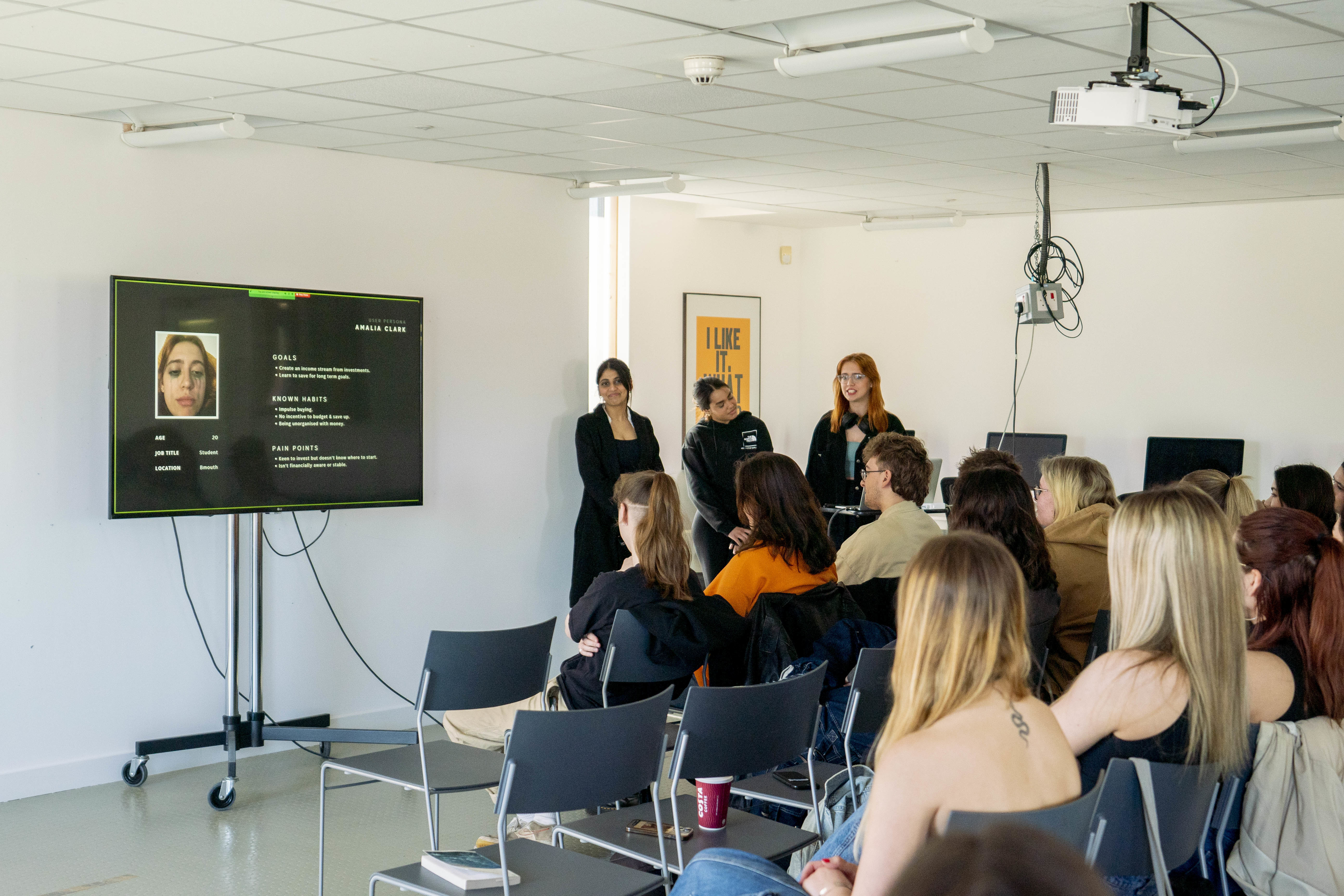 Three people stood at the front of a classroom giving a presentation with a monitor standing to their left.