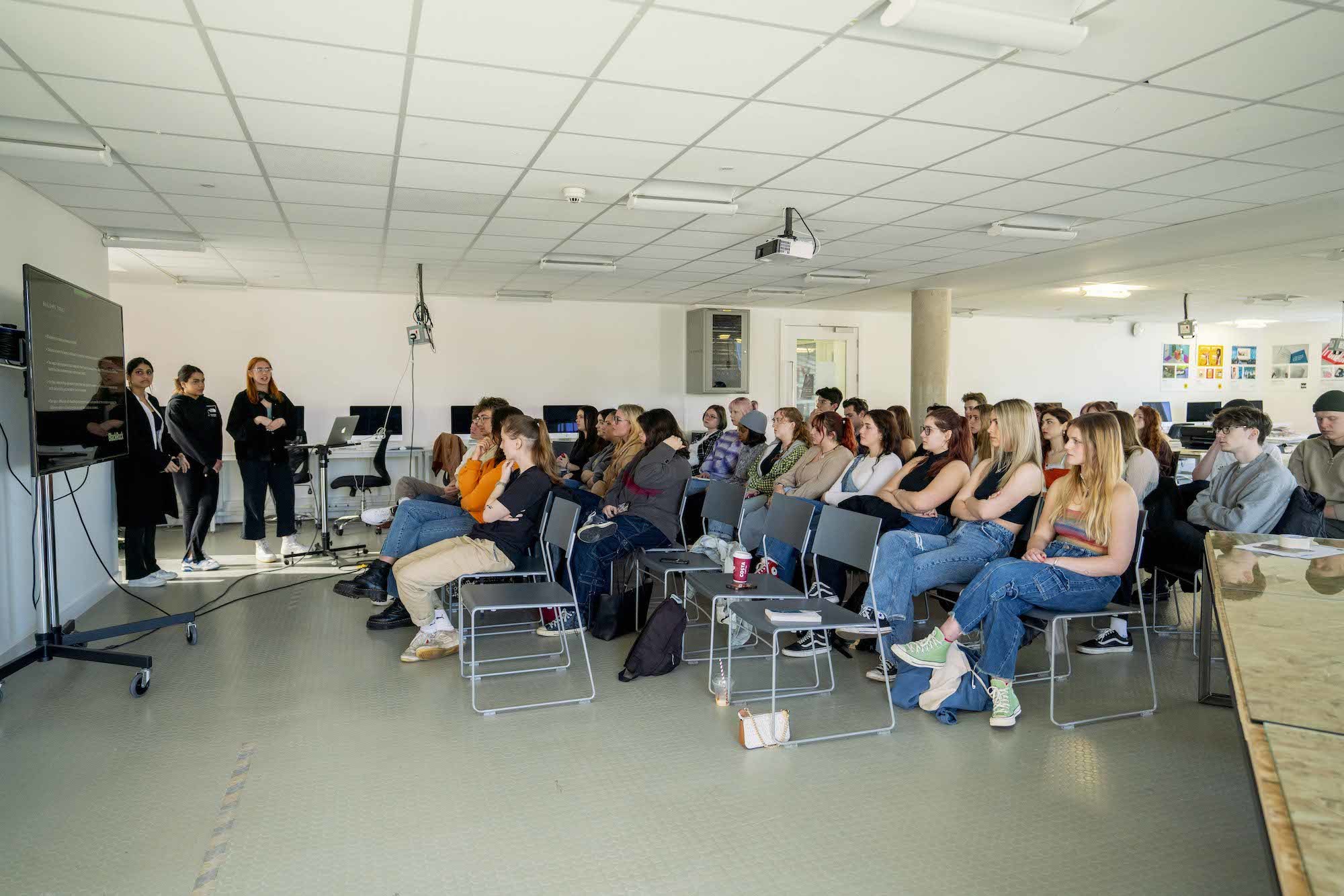 Three people deliver a presentation in front of a large group of people sitting in rows of seats in front of them.