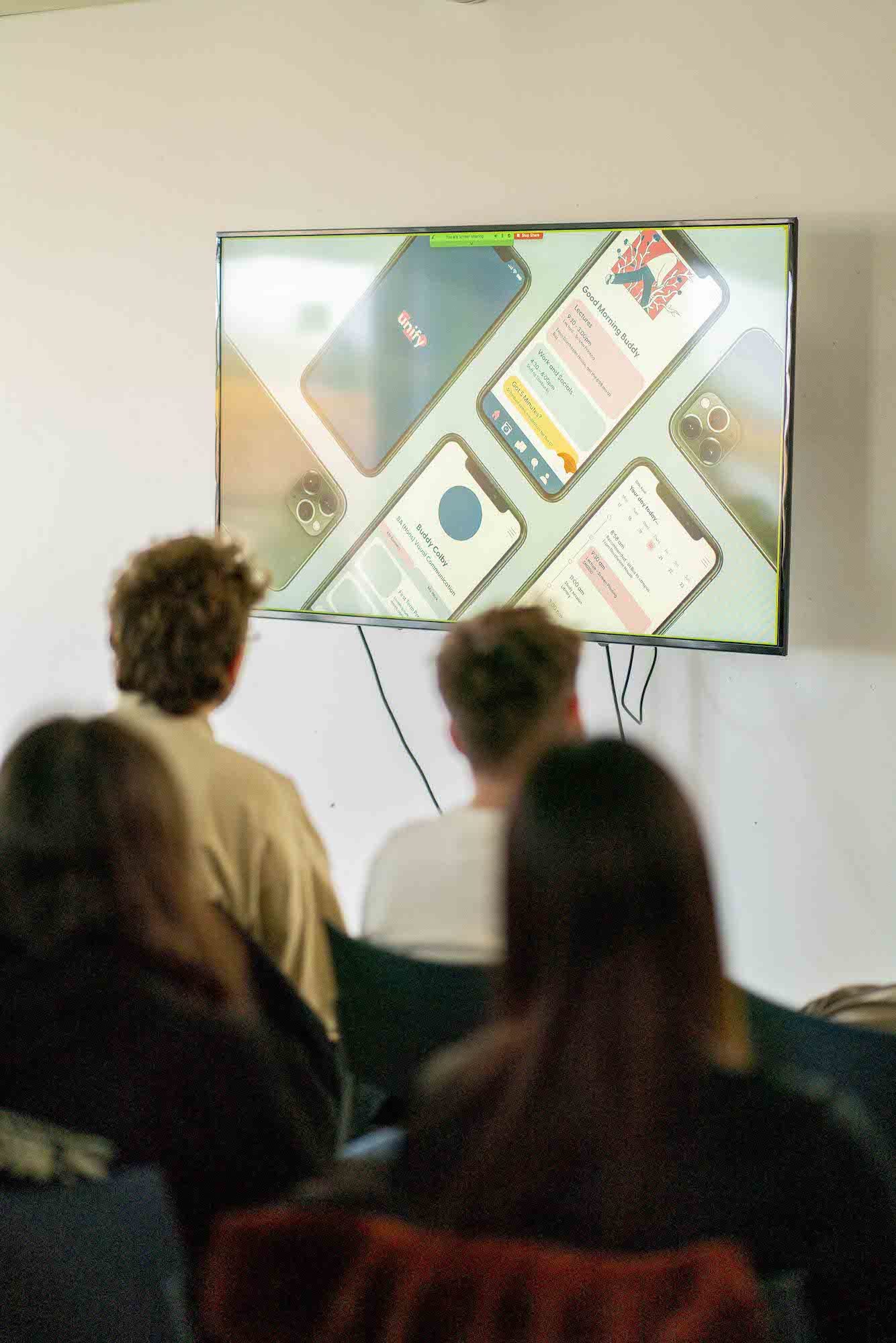 A monitor showing several mobile phone mock-ups, with people sitting on chairs in the foreground.