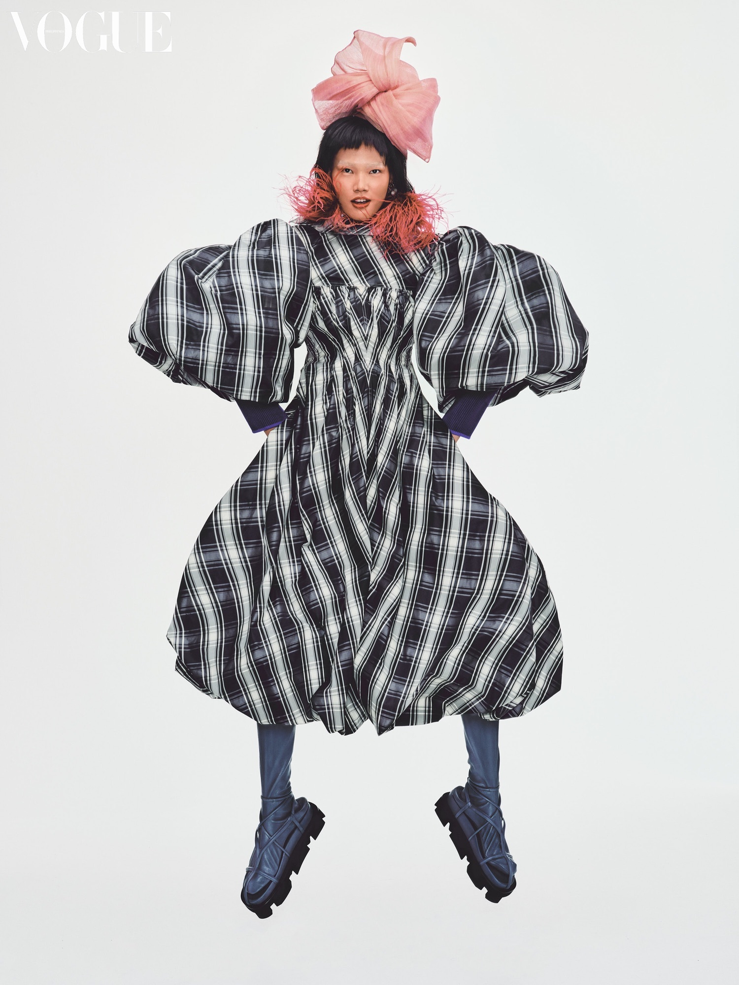 A model wearing a large black and white checkered dress, with heavy black shoes and pink headwear on a white background.