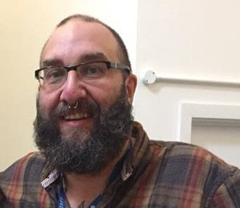 Bearded man with glasses wearing plaid shirt, smiling at camera in indoor setting with white door frame visible behind.