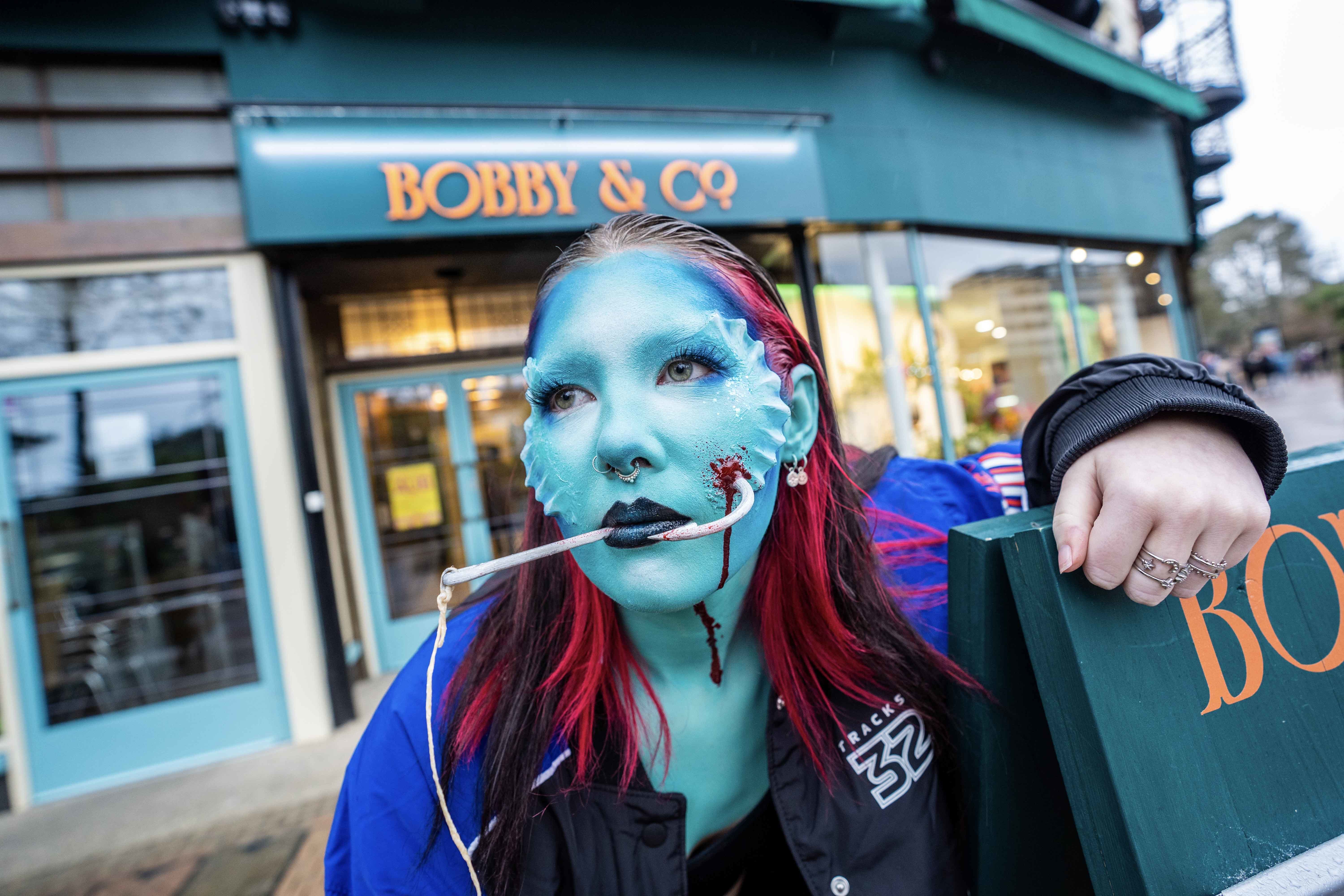 Student poses in front of a department store wearing facial prosthetics to show a fishing hook going through their cheek.