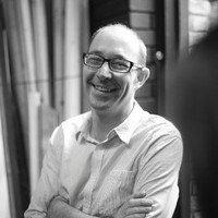 Bald man with glasses wearing light-coloured shirt, smiling whilst seated indoors in black and white photograph.