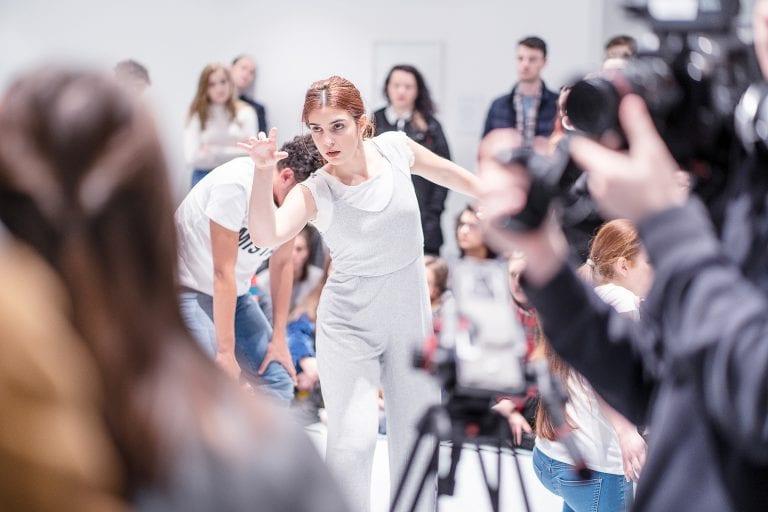A performer leans forward about to stretch out their arm. A crowd of people watch from around a gallery space.