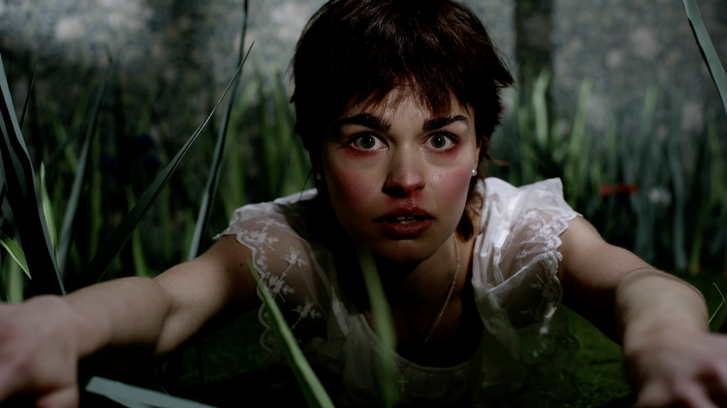 Young person with dark hair and wide eyes lies amongst tall green grass, wearing a white top, looking directly at camera.