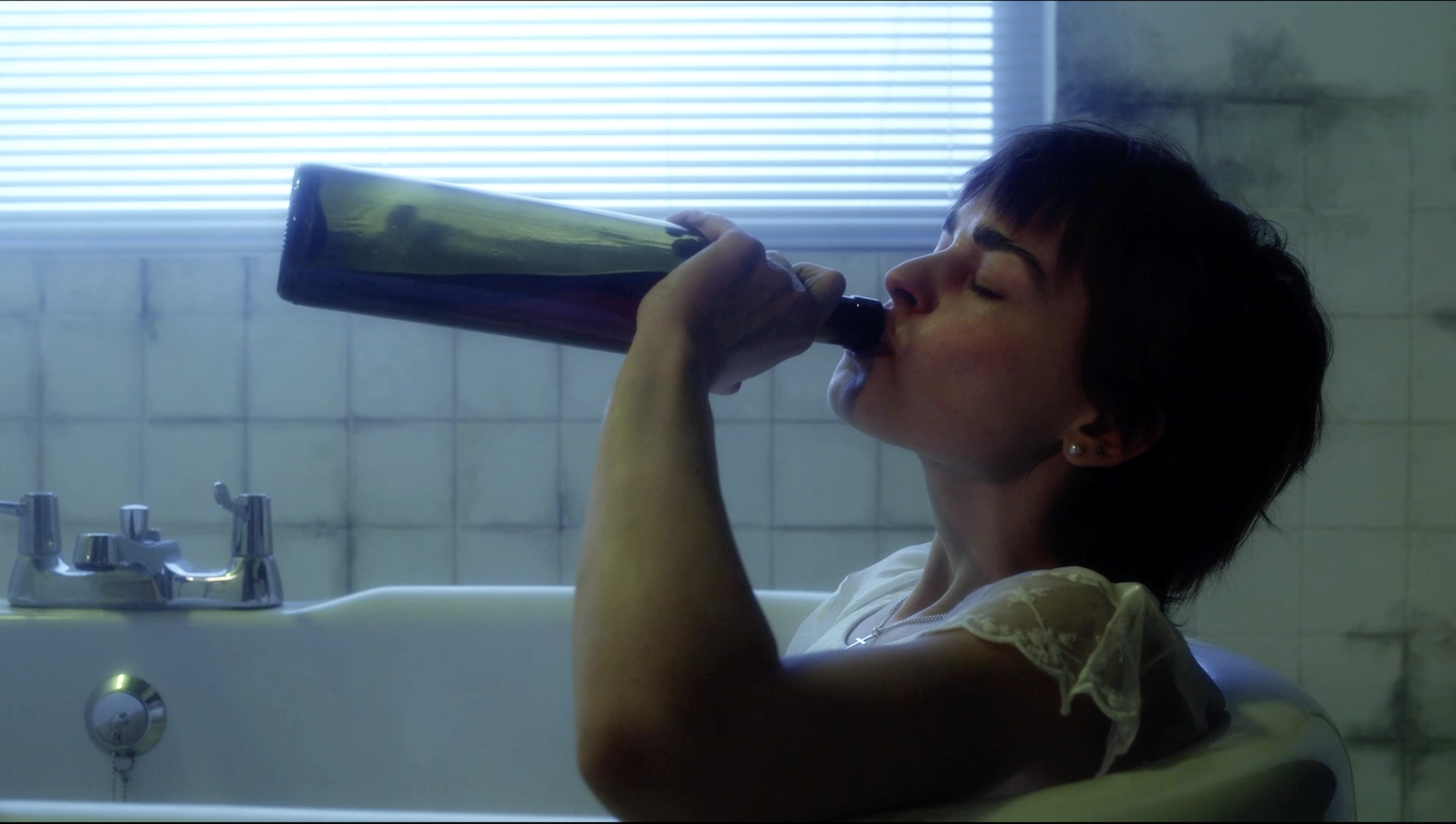 Person with dark hair drinking from green bottle in bathroom with white tiles and window blinds in background.