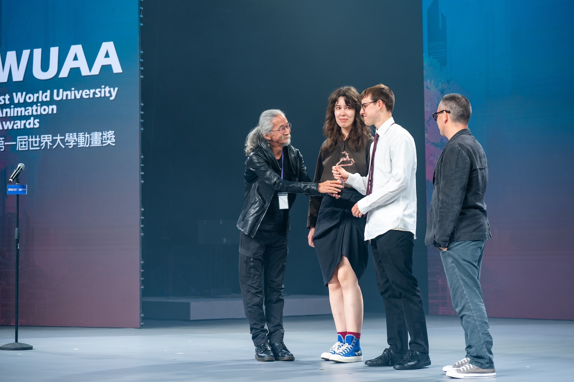 Award ceremony on stage with four people, one presenting trophy to M. Lebedeva and O. Mottram whilst fourth person watches. Blue backdrop displays "WUAA" text.