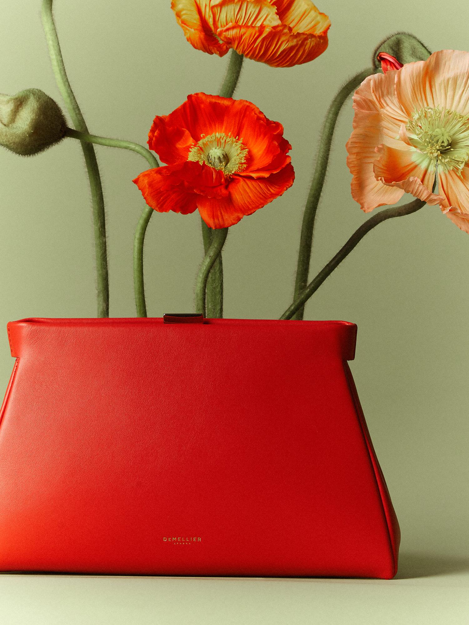 A red handbag placed on a smooth surface, with large, vibrant flowers of orange and peach emerging from it. The background is a muted green, highlighting the contrasts of the handbag and the floral arrangement.