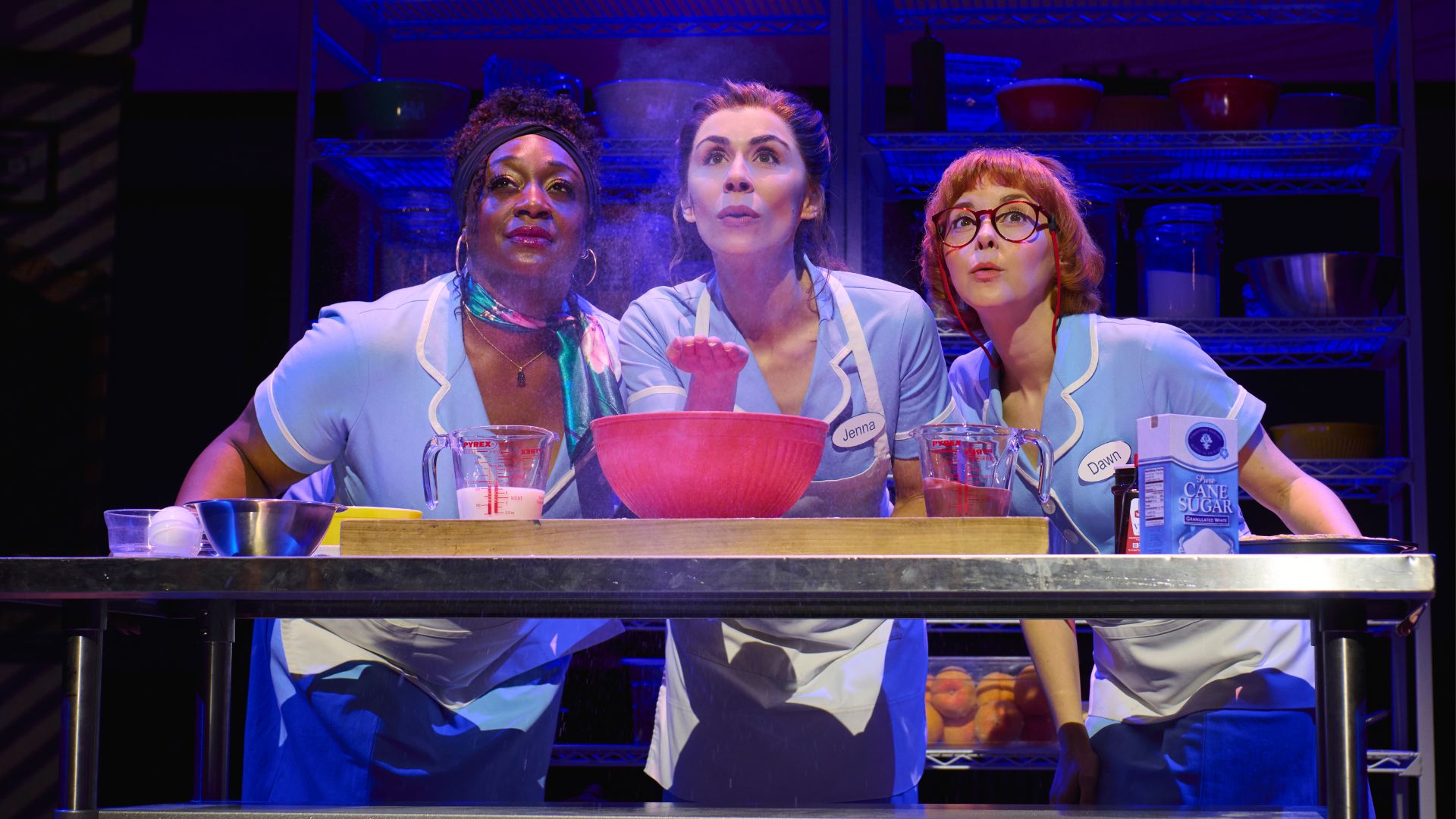 Three restaurant staff in blue uniforms and aprons stand behind a stainless-steel counter with bowls and kitchen utensils.