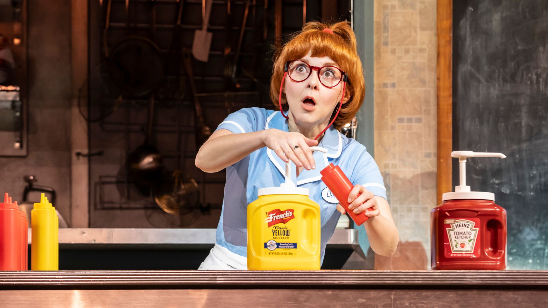 Actor E Hoskins portraying a waitress with red hair and glasses in a blue uniform holding a red condiment bottle behind a counter, with yellow and red squeeze bottles beside them.