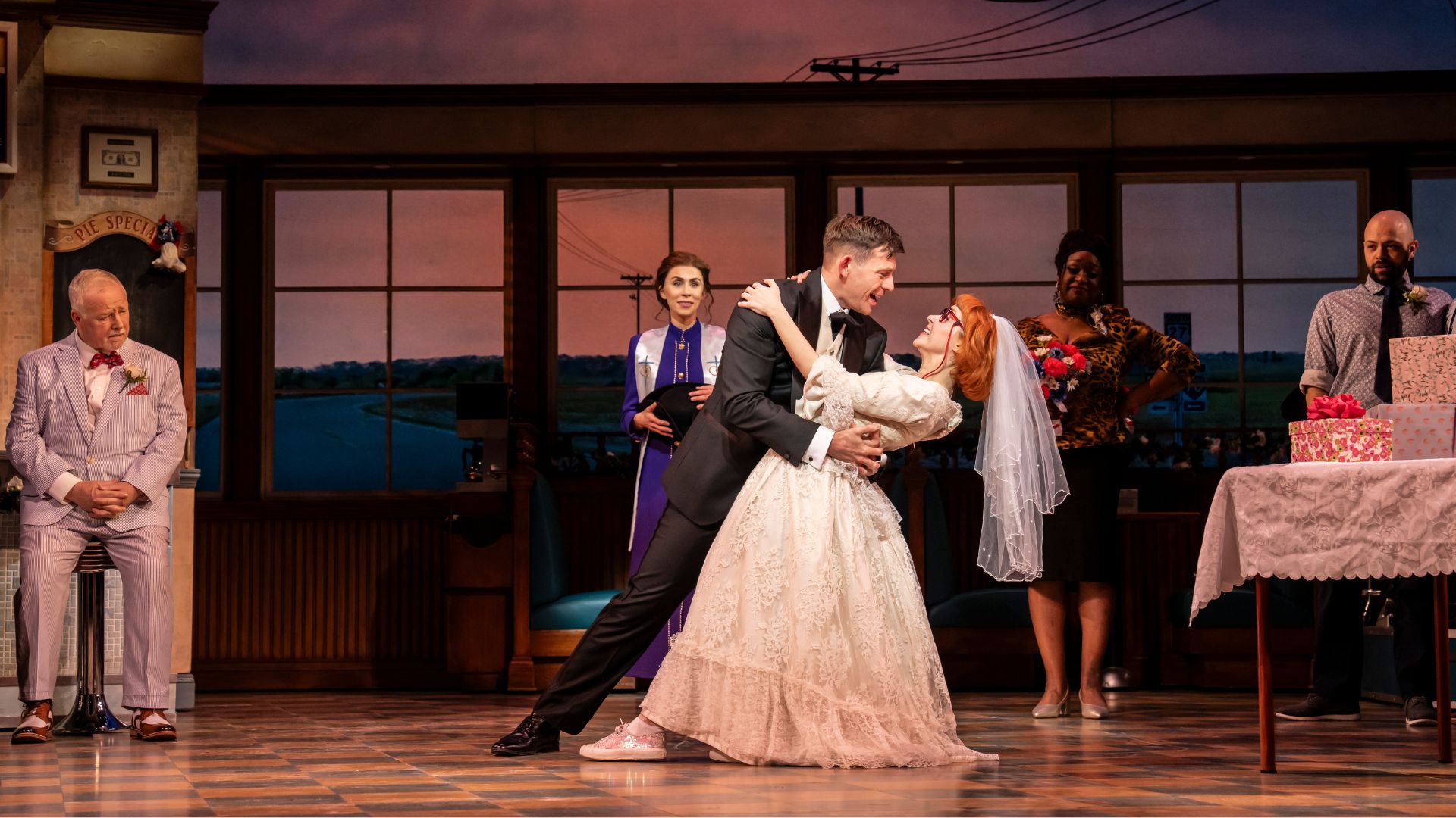A bride in a white wedding dress dances with a groom in a dark suit on a stage, with other guests standing beside a table in the background.