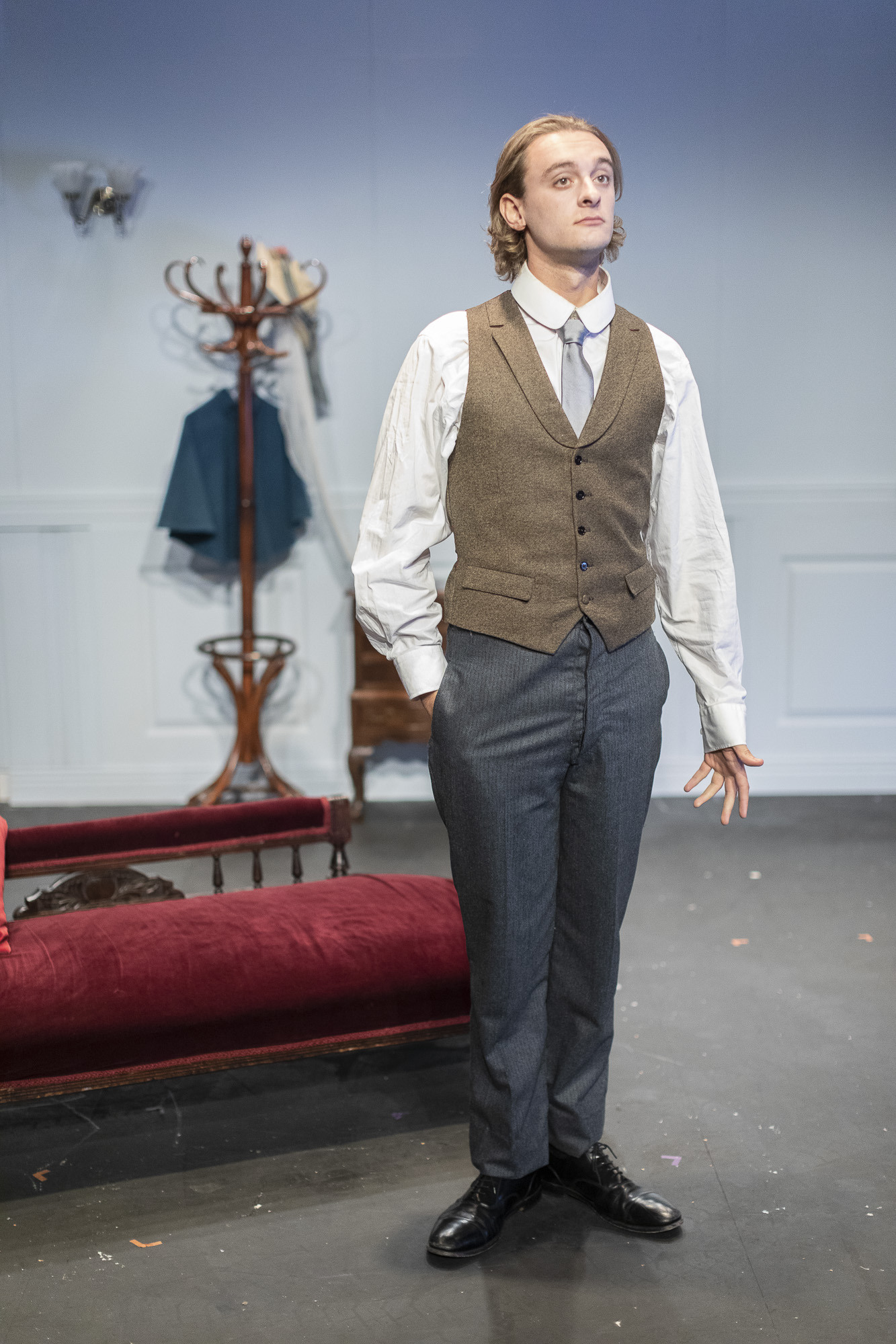 An actor on stage in a waistcoat standing on stage in front of a Roman couch.