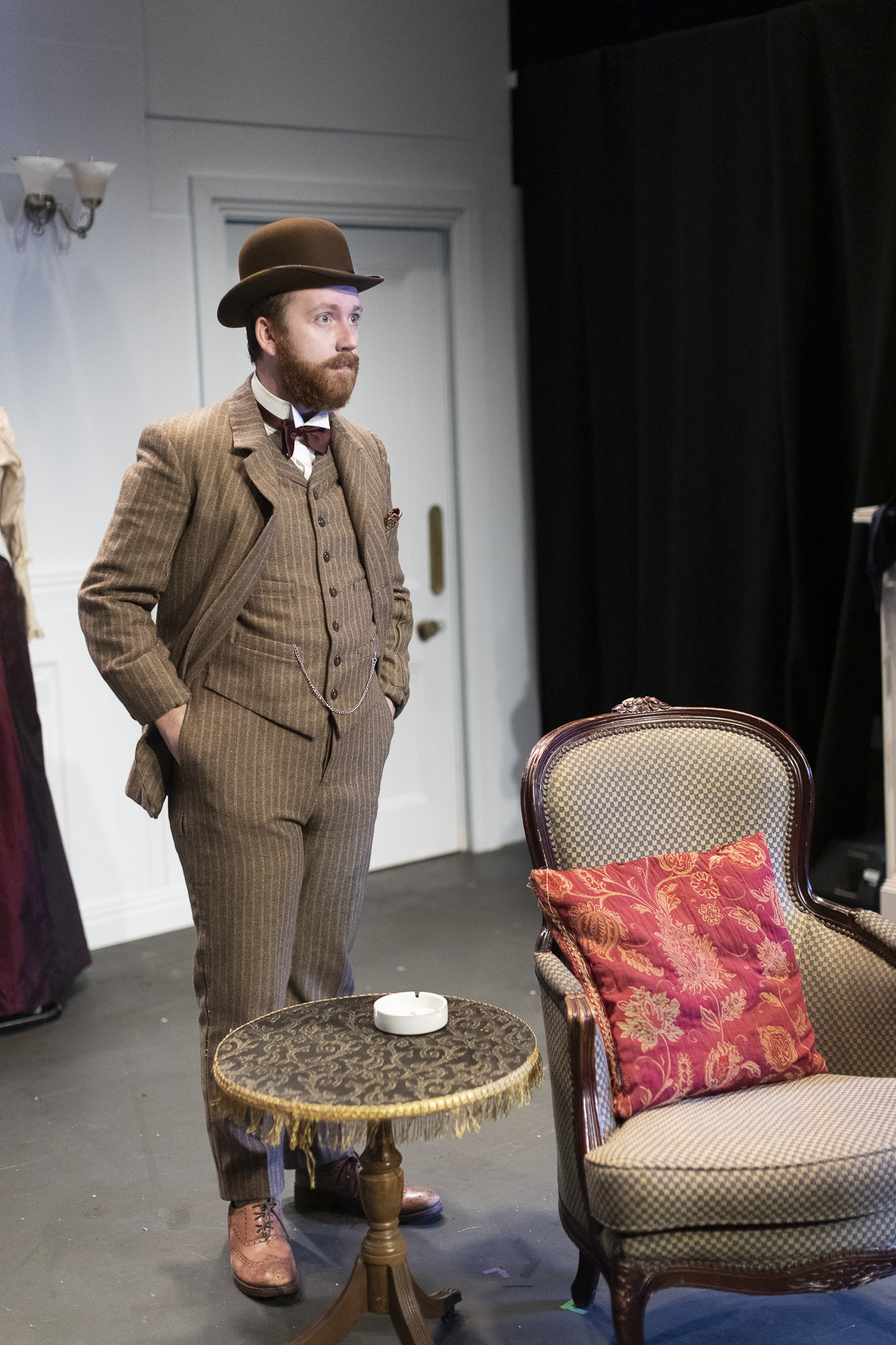 An actor on stage in costume – a pinstripe suit and bowler hat – standing behind an armchair and a tea table.