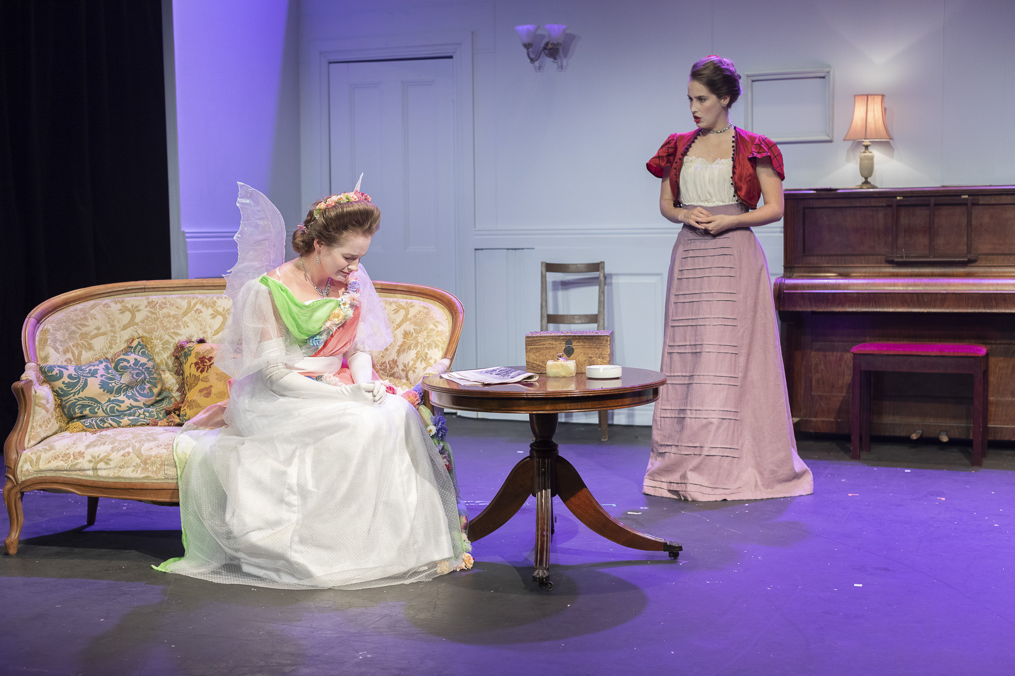 Two actors in costume in the middle of a performance. One actor is sitting on a sofa next to a coffee table, the other is standing next to an upright piano.
