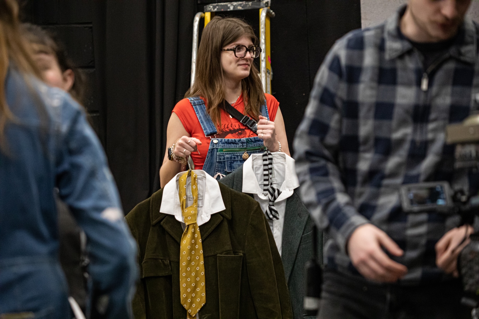 Woman with glasses in red shirt and denim dungarees holds up clothing items on hangers, with two people in background.