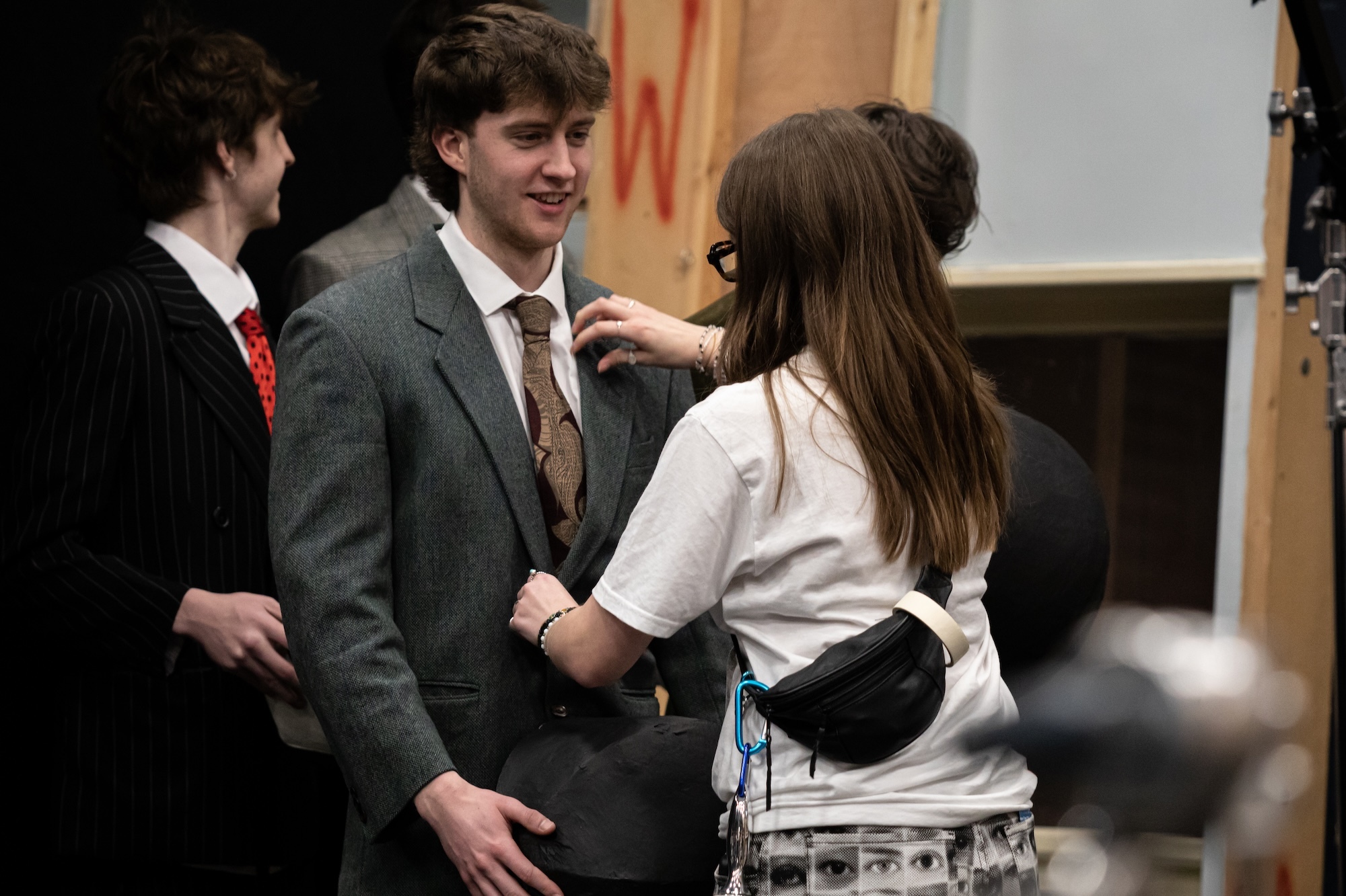 Young man in grey suit having his tie adjusted by woman in white shirt, with another person in dark clothing visible to the left.