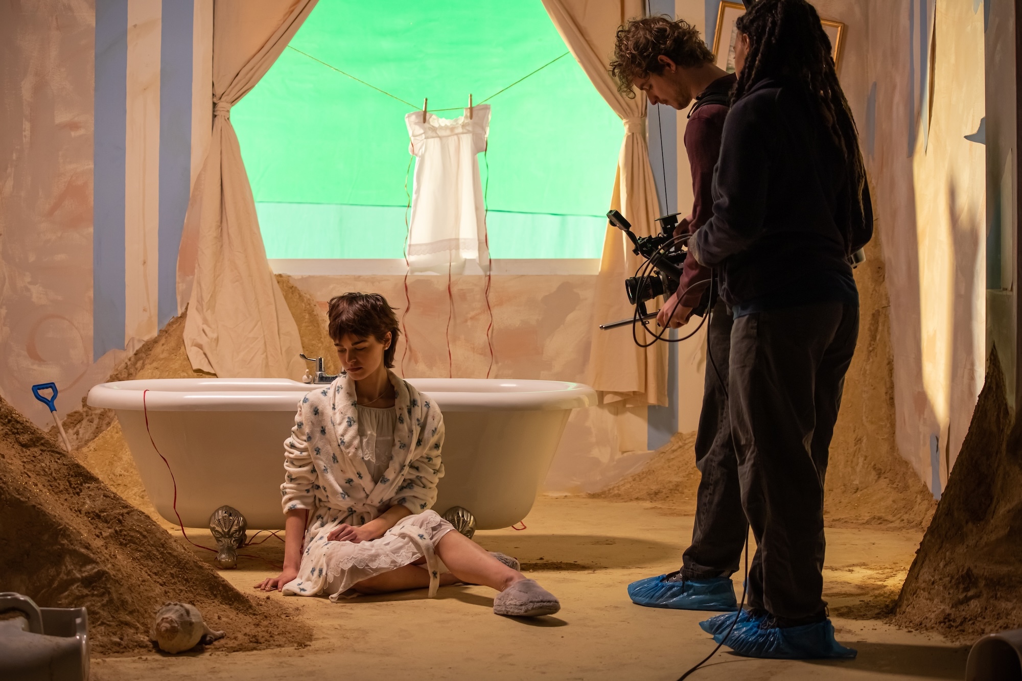Film set with cameraman recording actor in white robe sitting beside bathtub, green screen visible in background.