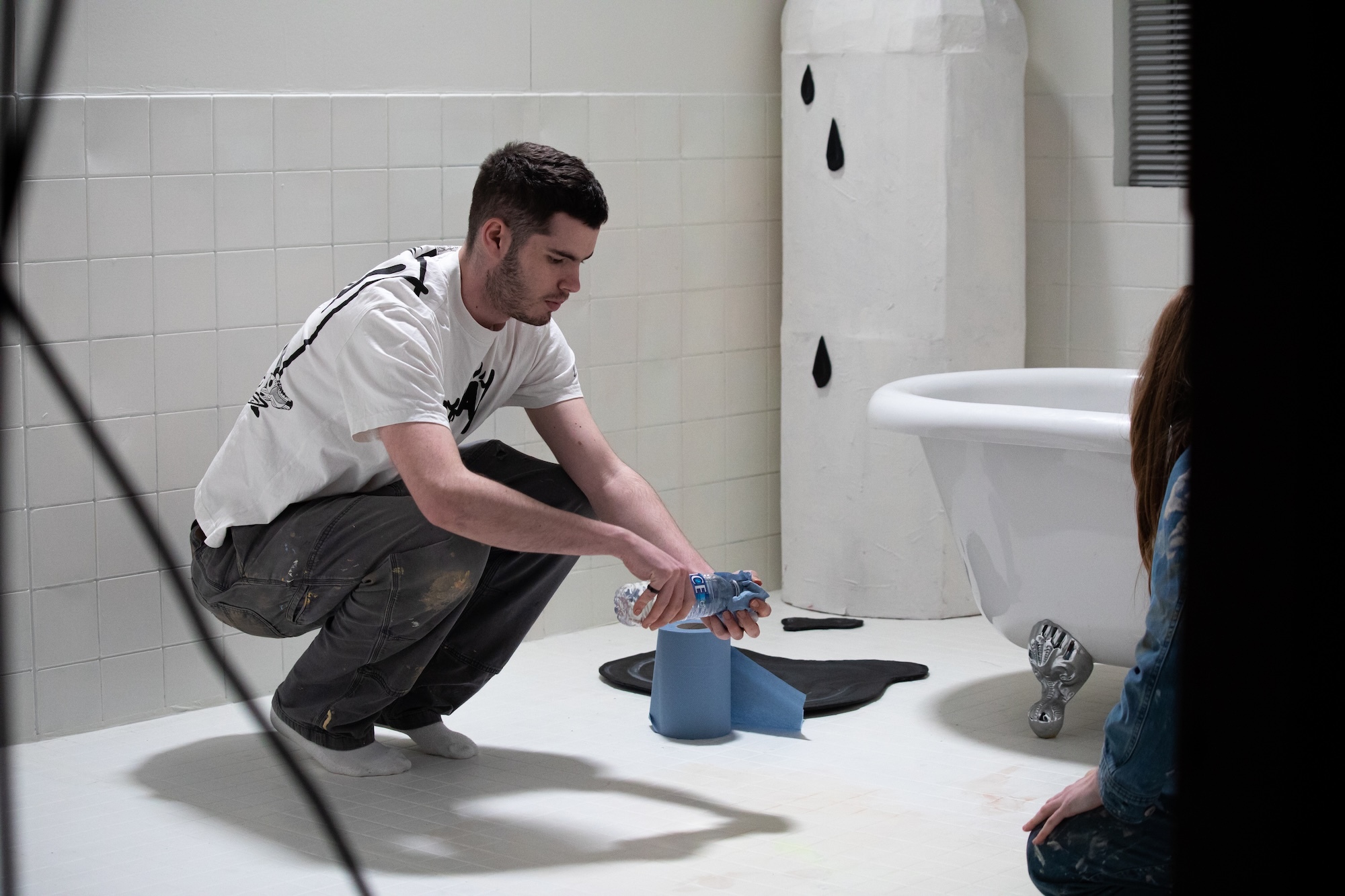 Man in white shirt crouching on white bathroom floor, placing blue container next to dark spill beneath leaking pipe.