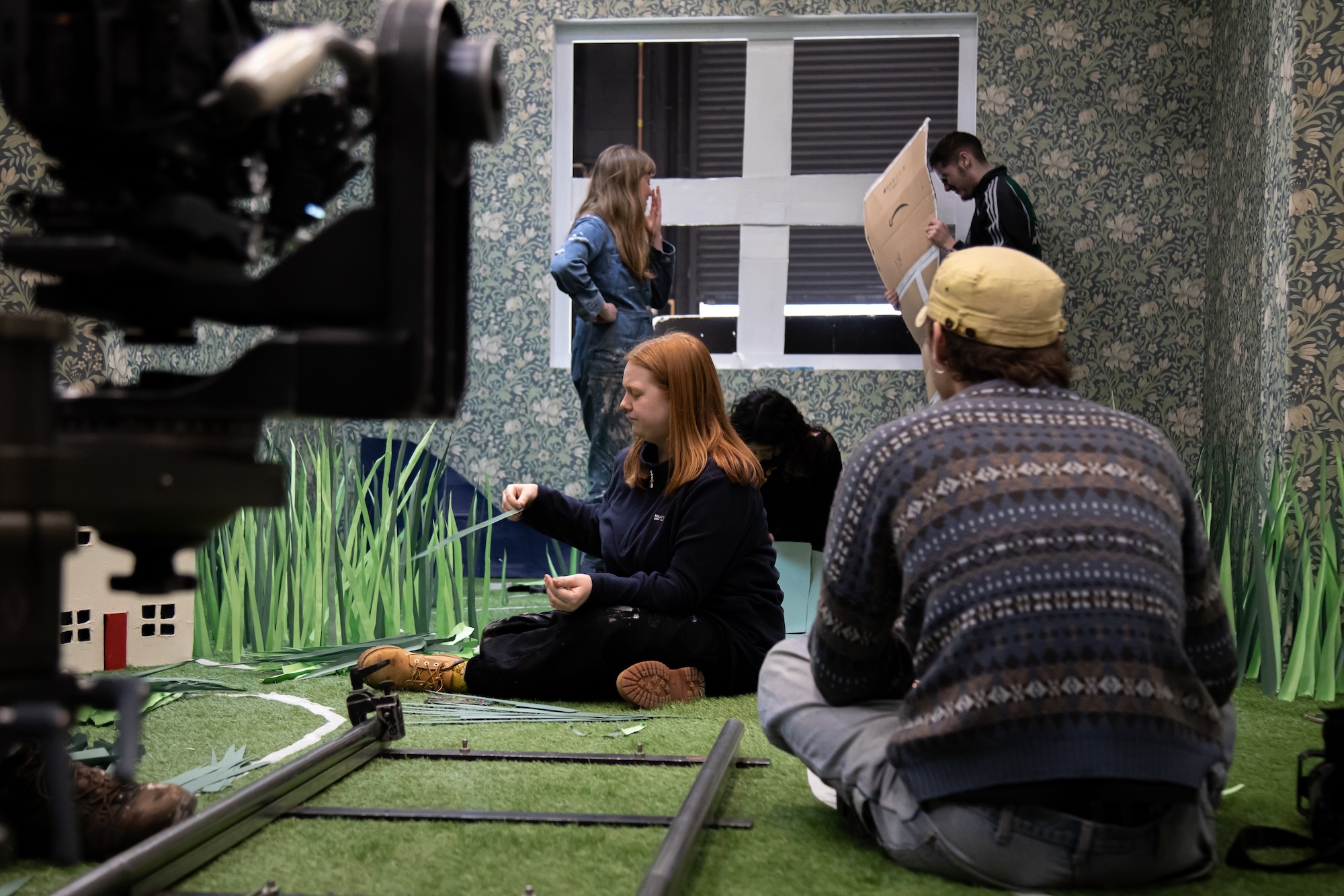 Film crew recording scene with actors on green set featuring miniature house, painted grass backdrop, and window prop.