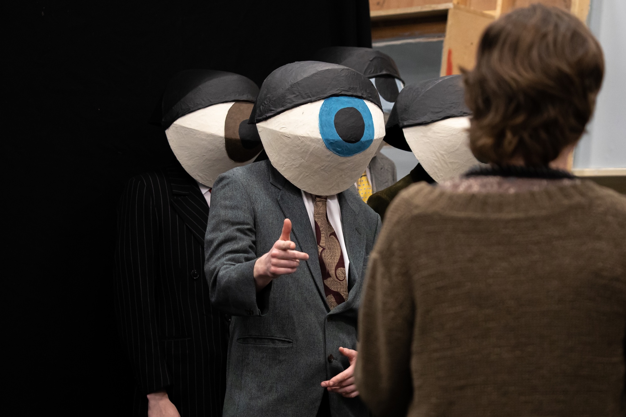 Person in grey suit wearing large white mask with blue centre circle and black eyebrows, speaking to seated individual.