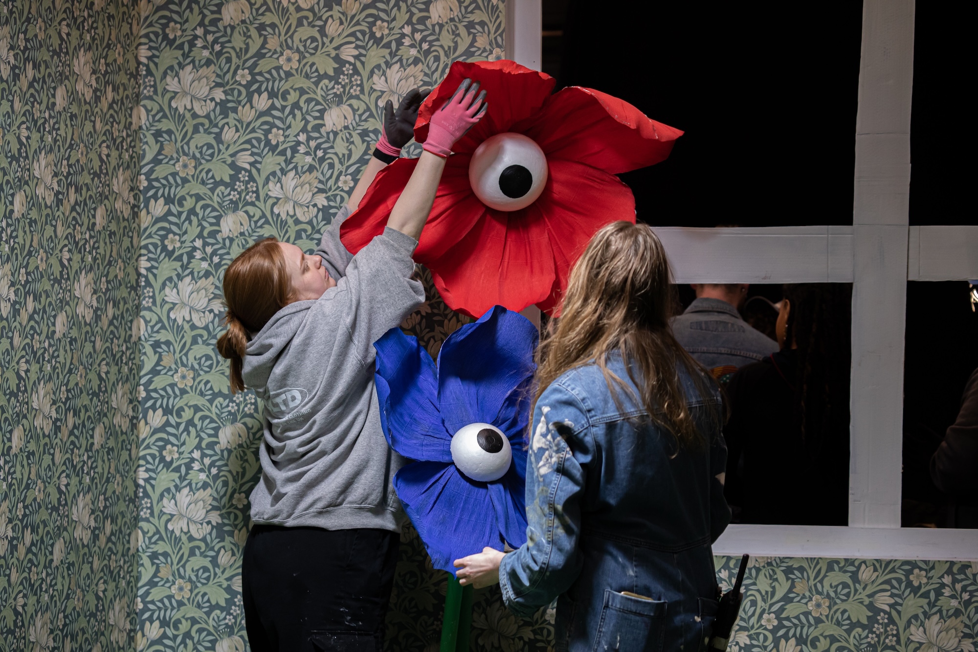 Two people holding large red and blue puppet heads with white circular eyes against patterned wallpaper and window frame.