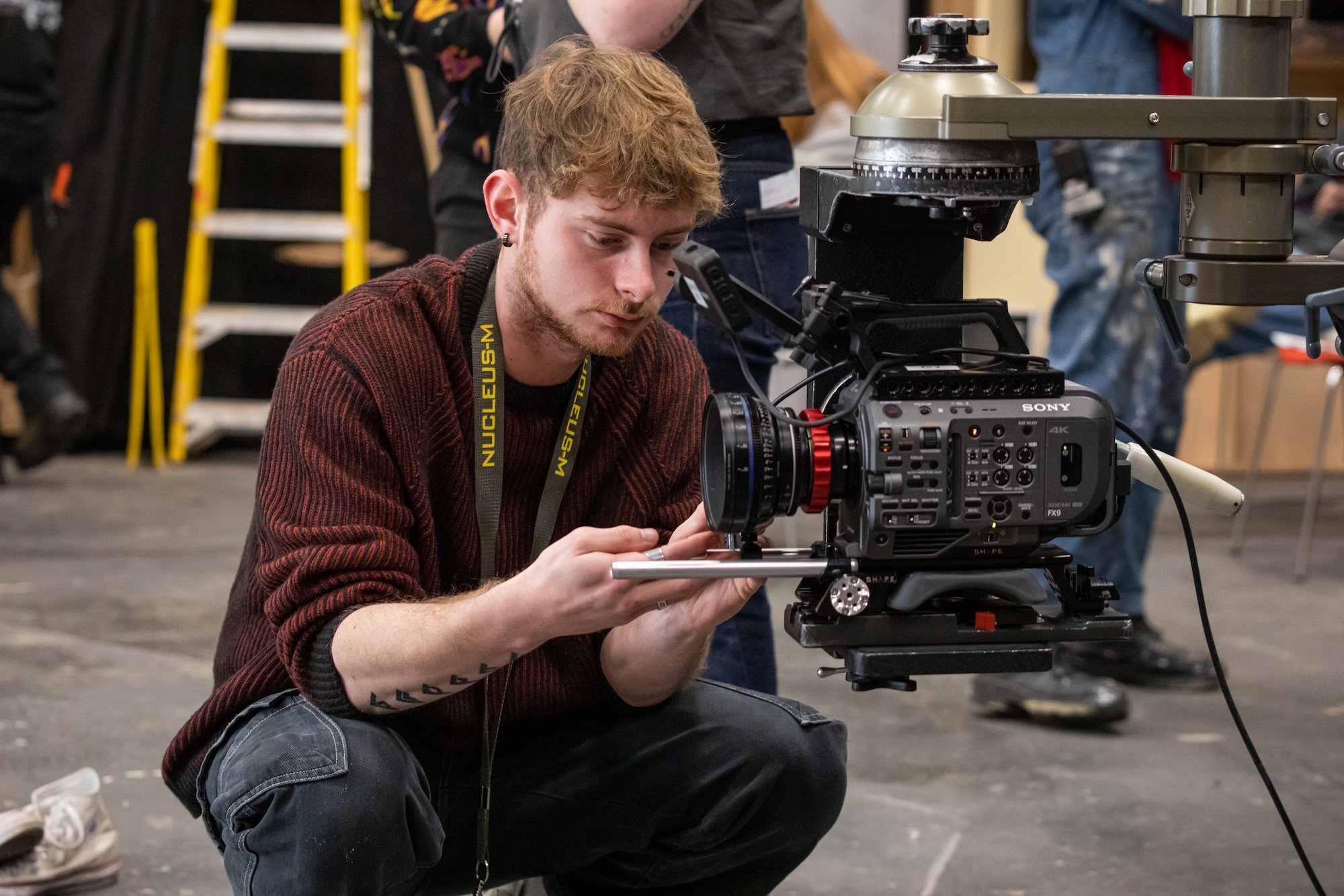 A BA (Hons) Film Production student crouching beside professional film camera on set, adjusting equipment with crew in background.