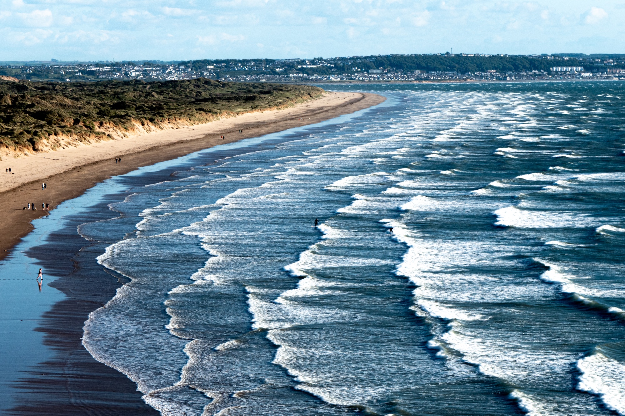A sweeping sandy beach with white surf rolling onto the shore, with coastal dunes and a distant town along the horizon.